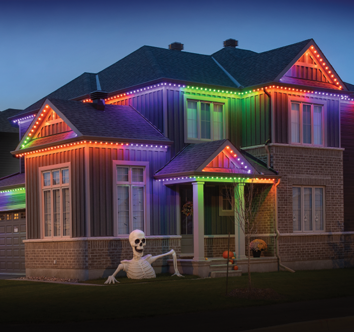 A two-story house decorated with colorful Halloween lights, a skeleton figure in front yard, pumpkins, and fall decorations.