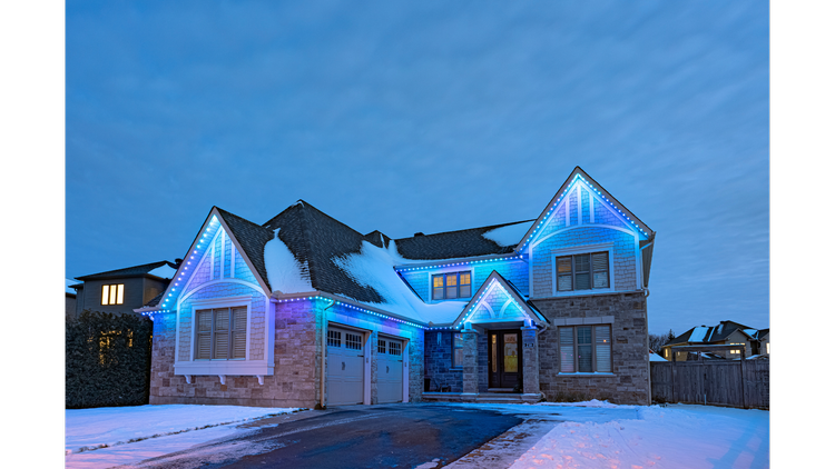 A two-story house illuminated with blue string lights, snow on the roof and ground, and a paved driveway leading to a garage during twilight.