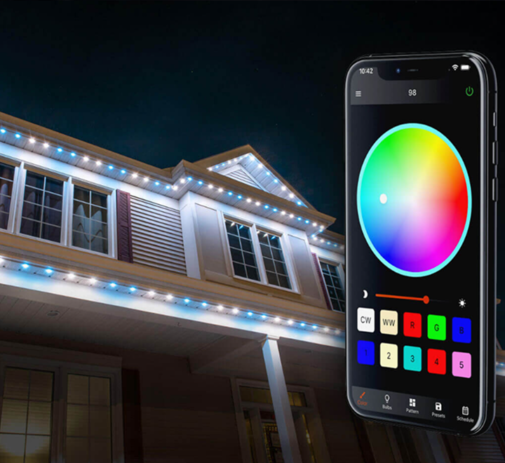 A smartphone displaying a color wheel app with controls, positioned in front of a house decorated with multicolored Christmas lights at night.