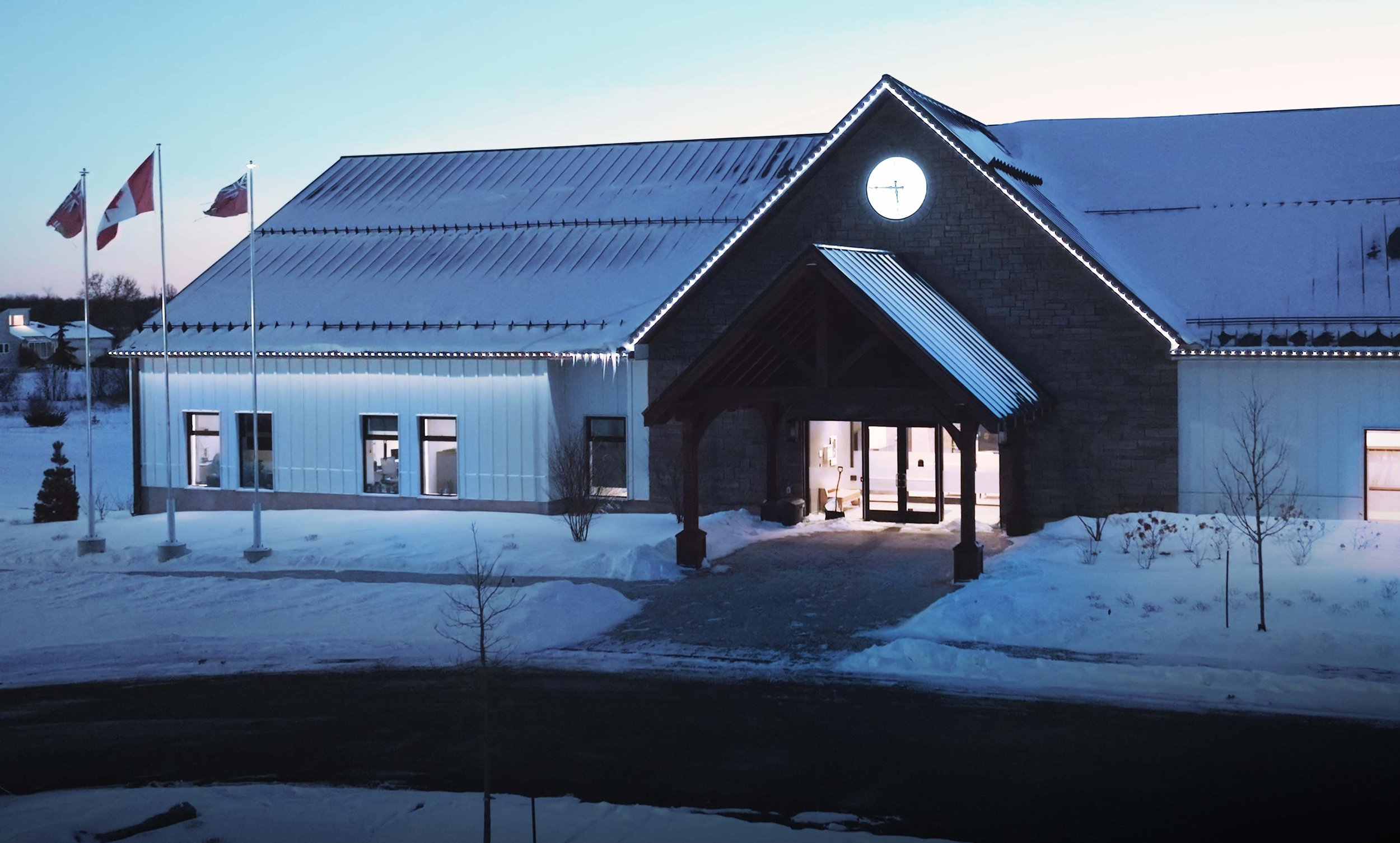 A snow-covered building with a clock on the upper gable, lit up with holiday lights along the roofline and around the entrance, during dusk or early evening.
