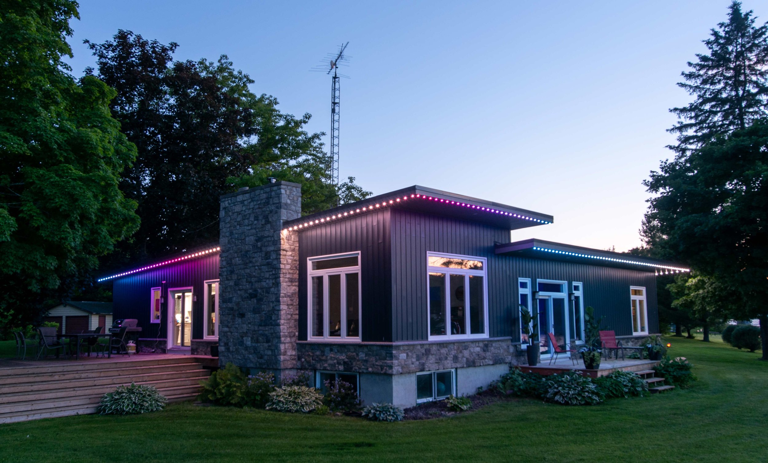 Modern house with dark siding, large windows, stone chimney, outdoor patio with chairs, and purple and blue LED string lights at dusk, surrounded by trees and a well-maintained lawn.