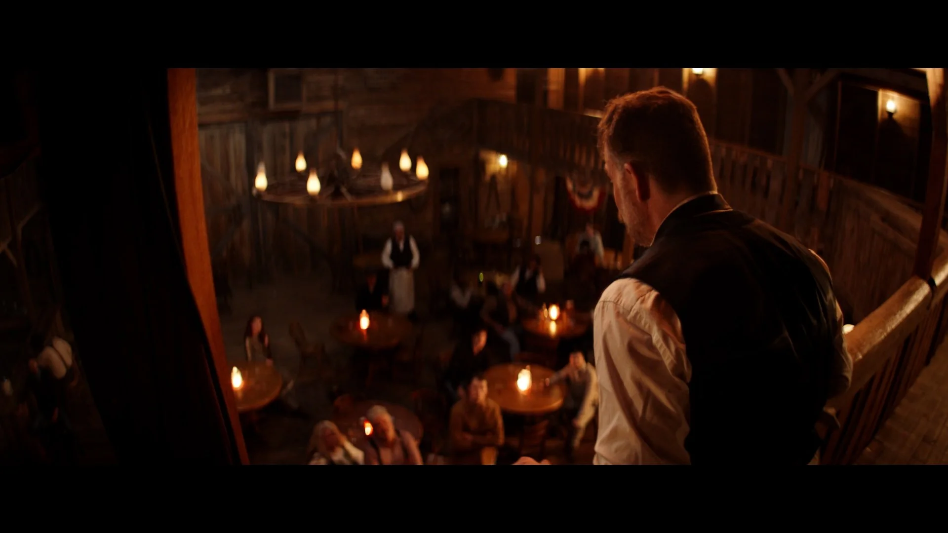 A man with a beard and wearing a vest, standing on a staircase balcony overlooking a rustic tavern filled with people, warm candle lighting, wooden interior, and a chandelier.
