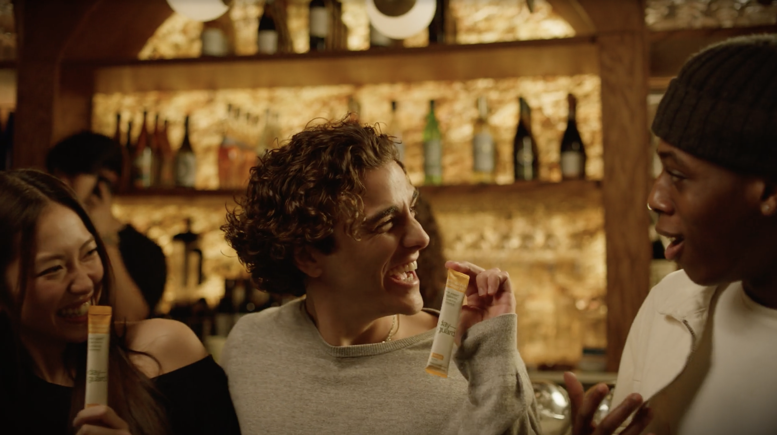 Four friends in a bar are smiling and chatting, with a woman holding a packet of snacks.