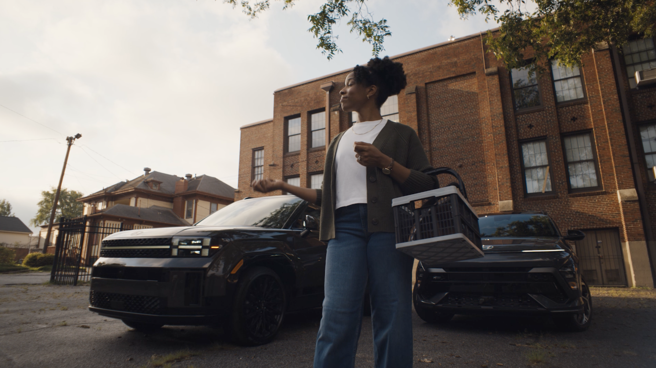 A woman walking outdoors near a Hyundai, carrying a laundry basket, with a brick apartment building in the background.