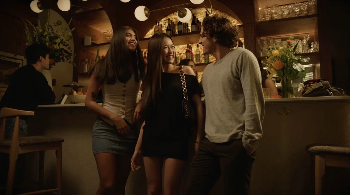 Three friends, two women and one man, are happily chatting and smiling at each other in a dimly lit bar or restaurant with a warm, cozy atmosphere. A bar counter with bottles and flowers is visible in the background.