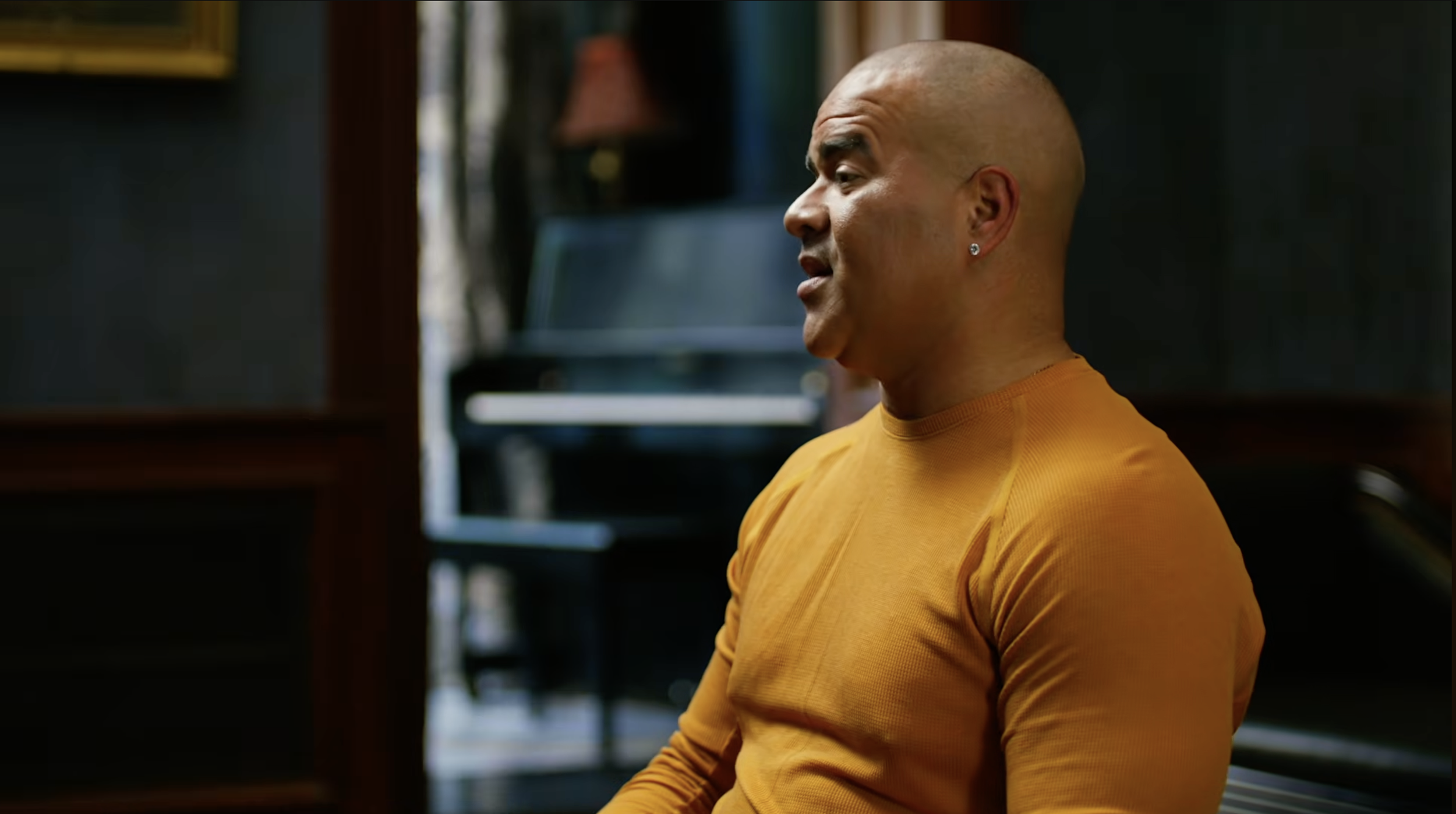 Chris Jackson with a shaved head wearing a yellow shirt, sitting indoors near a piano with a bookshelf in the background.