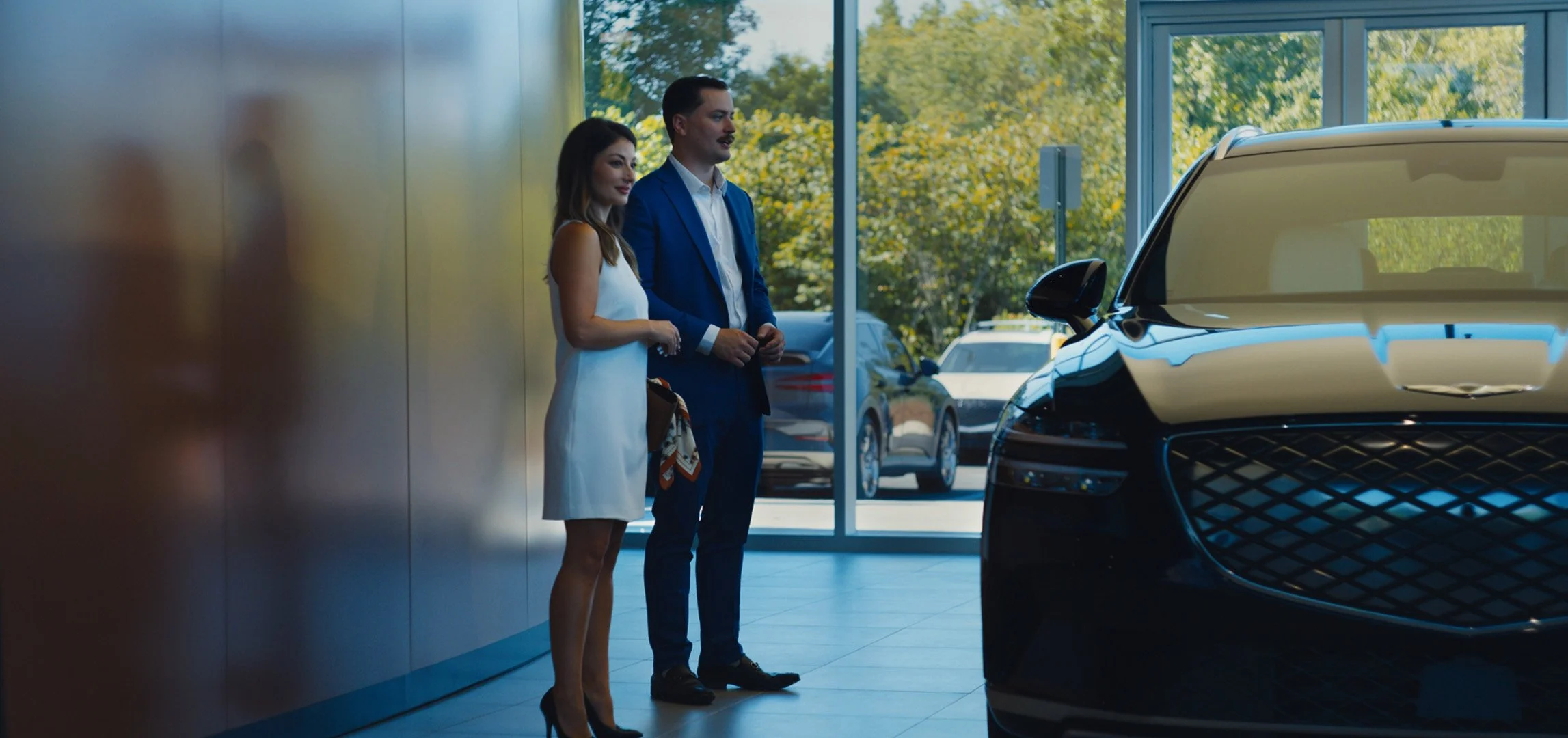 A man in a blue suit and a woman in a white dress standing inside a car dealership near a black Genesis, with large glass windows showing trees outside.