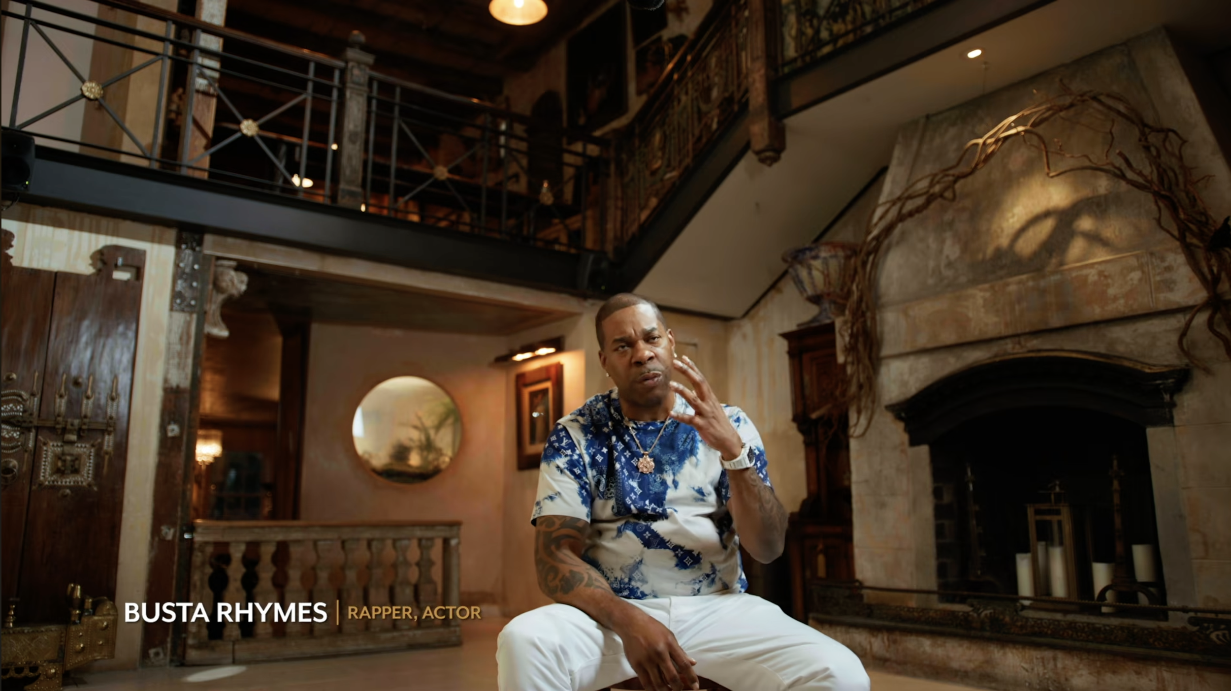 Busta Rhymes sitting in a spacious room with rustic decor, including a large stone fireplace, wooden beams, and ornate railings, with a thoughtful expression and his hand near his face.