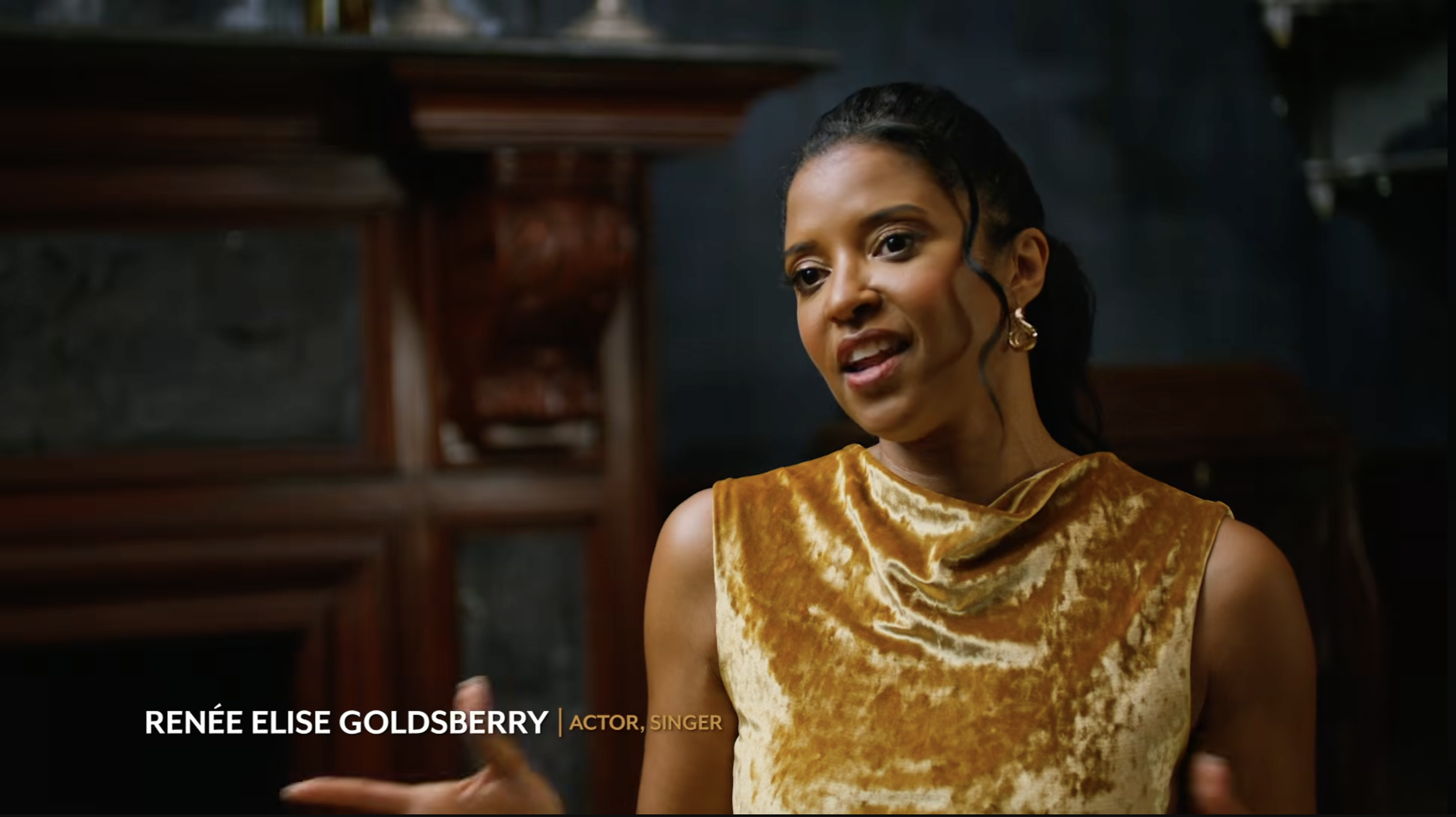 Renée Elise Goldsberry with dark hair in a ponytail is wearing a yellow velvet sleeveless top and gold earrings, standing in front of a dark background with wooden furniture visible.