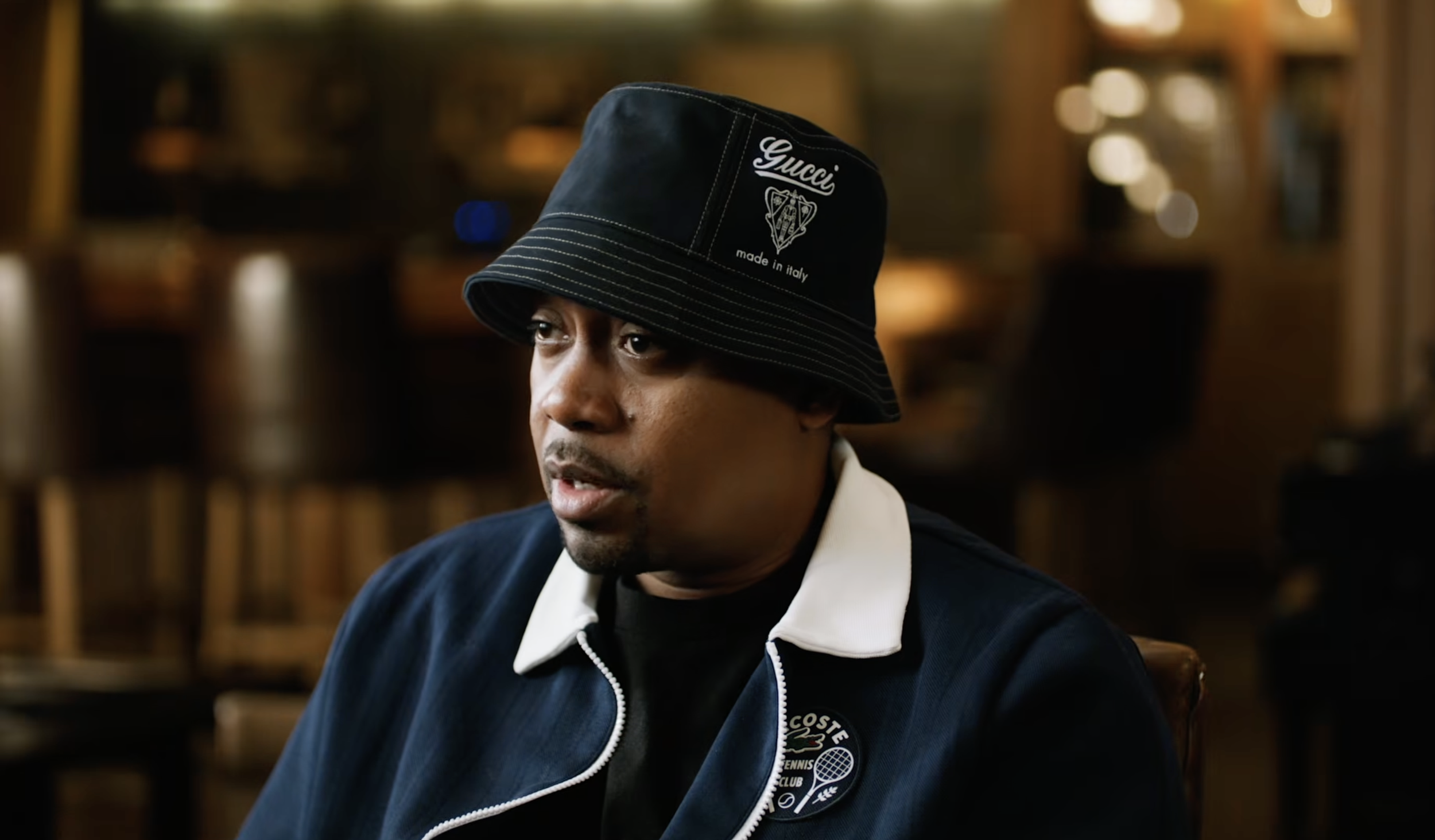 Nas wearing a black bucket hat with white text and graphics, a navy jacket, and a white shirt.
