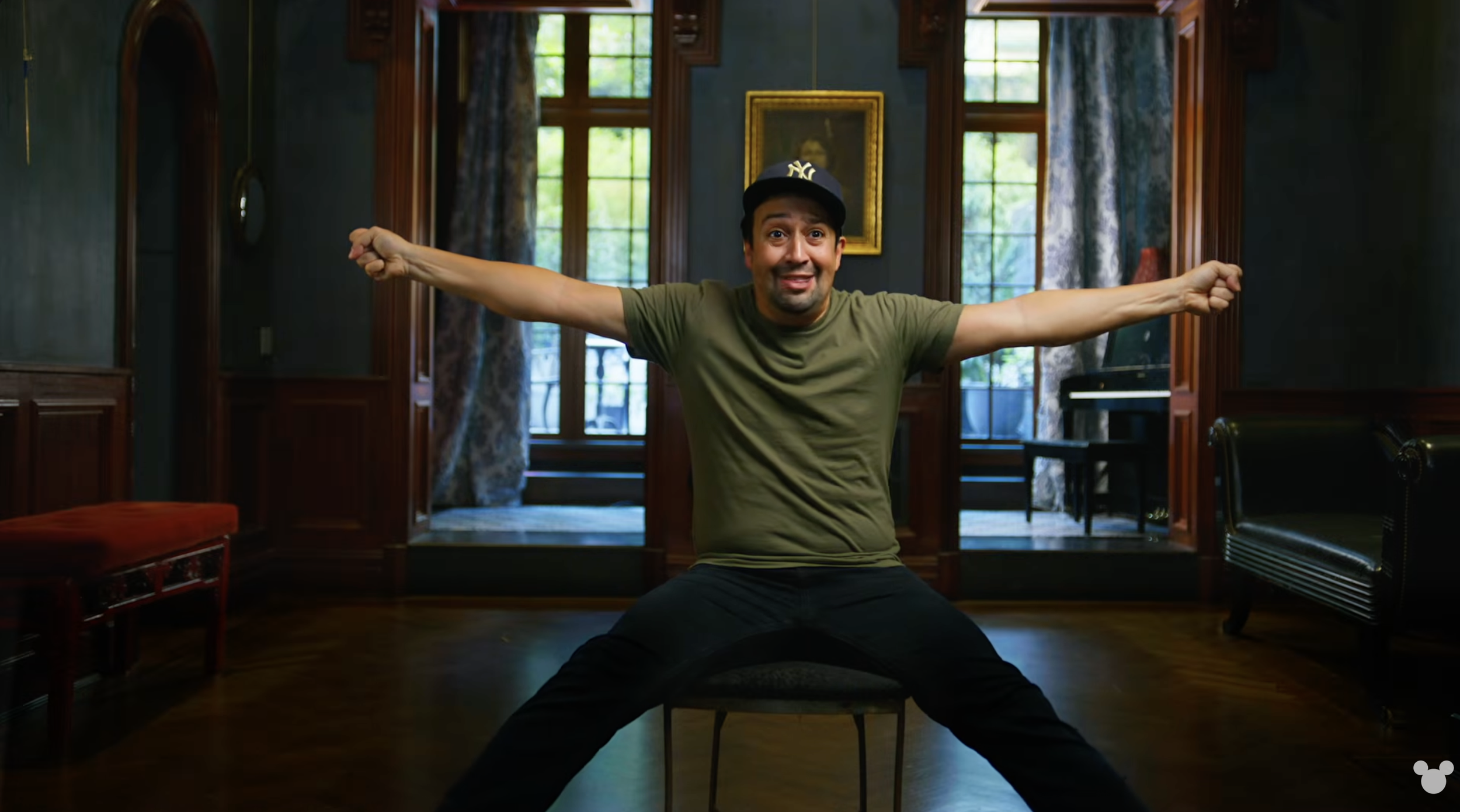 Lin-Manuel Miranda seated cross-legged with arms outstretched, smiling in a room with large windows, wooden trim, and a piano in the background.