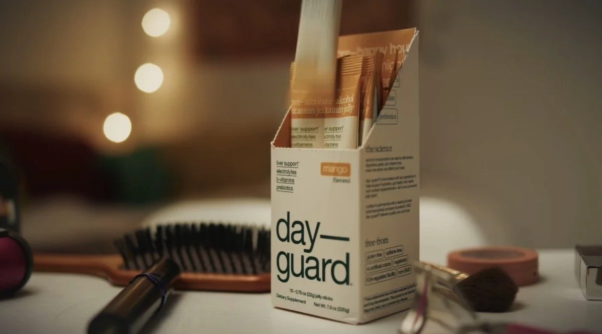 A box of dietary supplement sticks labeled 'day—guard' with a mango flavor on a table, surrounded by a makeup brush, hairbrush, and other beauty products.