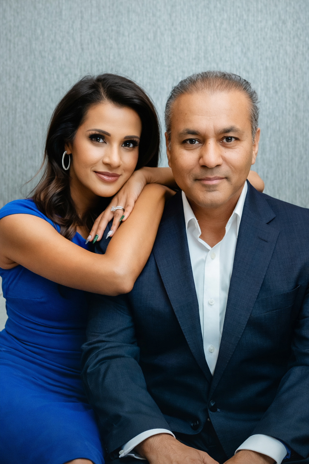 A young woman with dark hair wearing a blue dress and earrings, and an older man with graying hair in a suit and white shirt, posing close together with the woman resting her hand on the man's shoulder, against a light background.