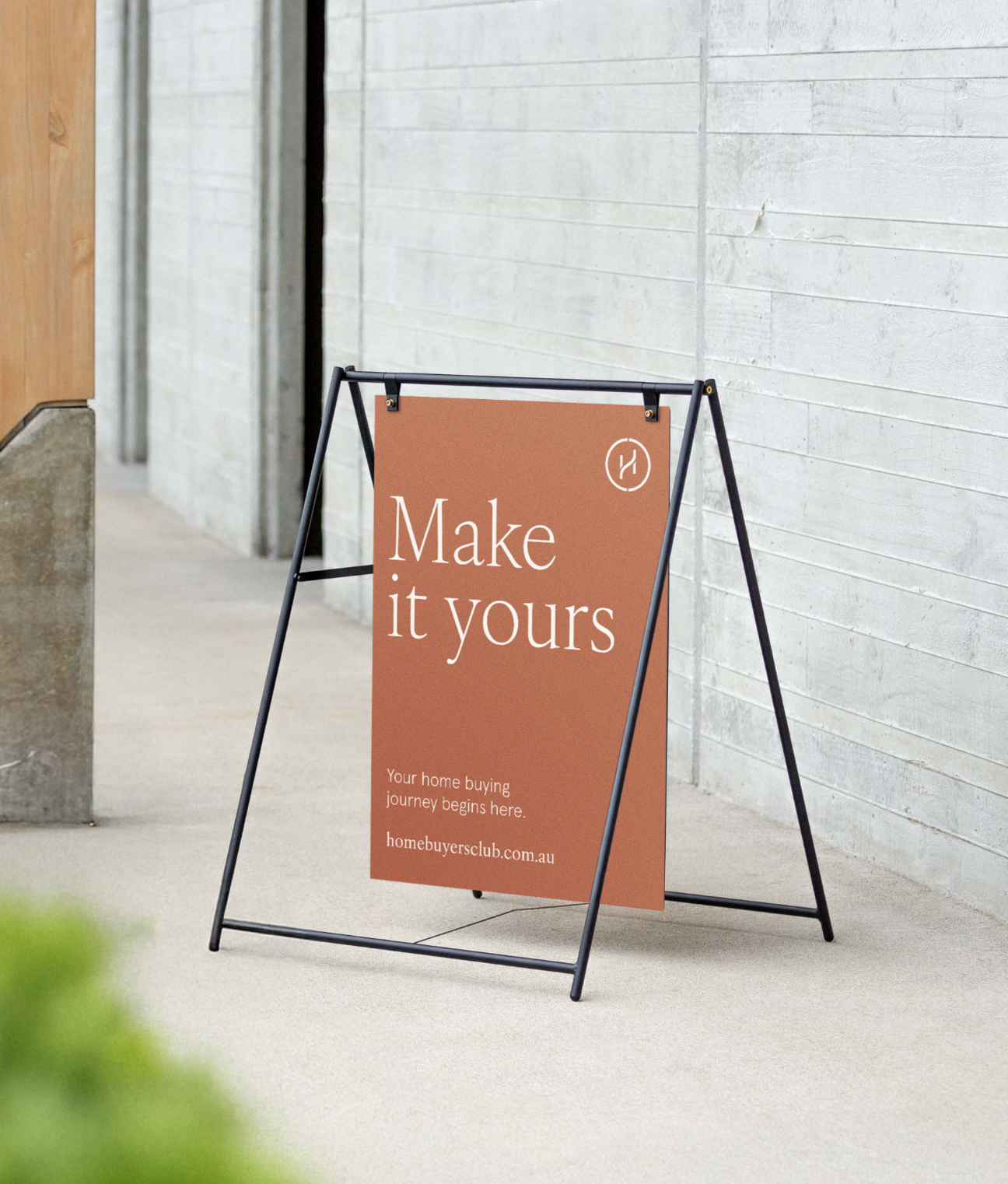 A sidewalk sign with a brown background and white text that reads "Make it yours." Underneath, it says "Your home buying journey begins here," and provides a website link to homebuyersclub.com.au. The sign is set against a concrete wall outside a building.