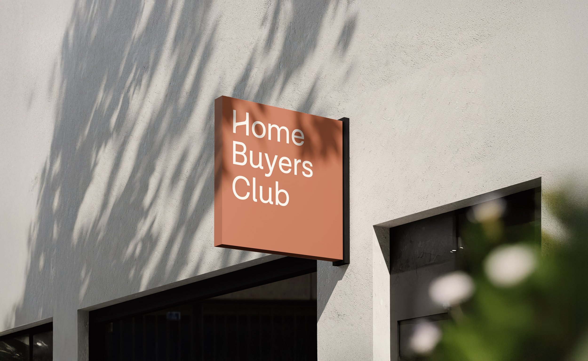 A sign on a white building reads 'Home Buyers Club' in white text on a pink background, with shadows of palm leaves cast on the wall.