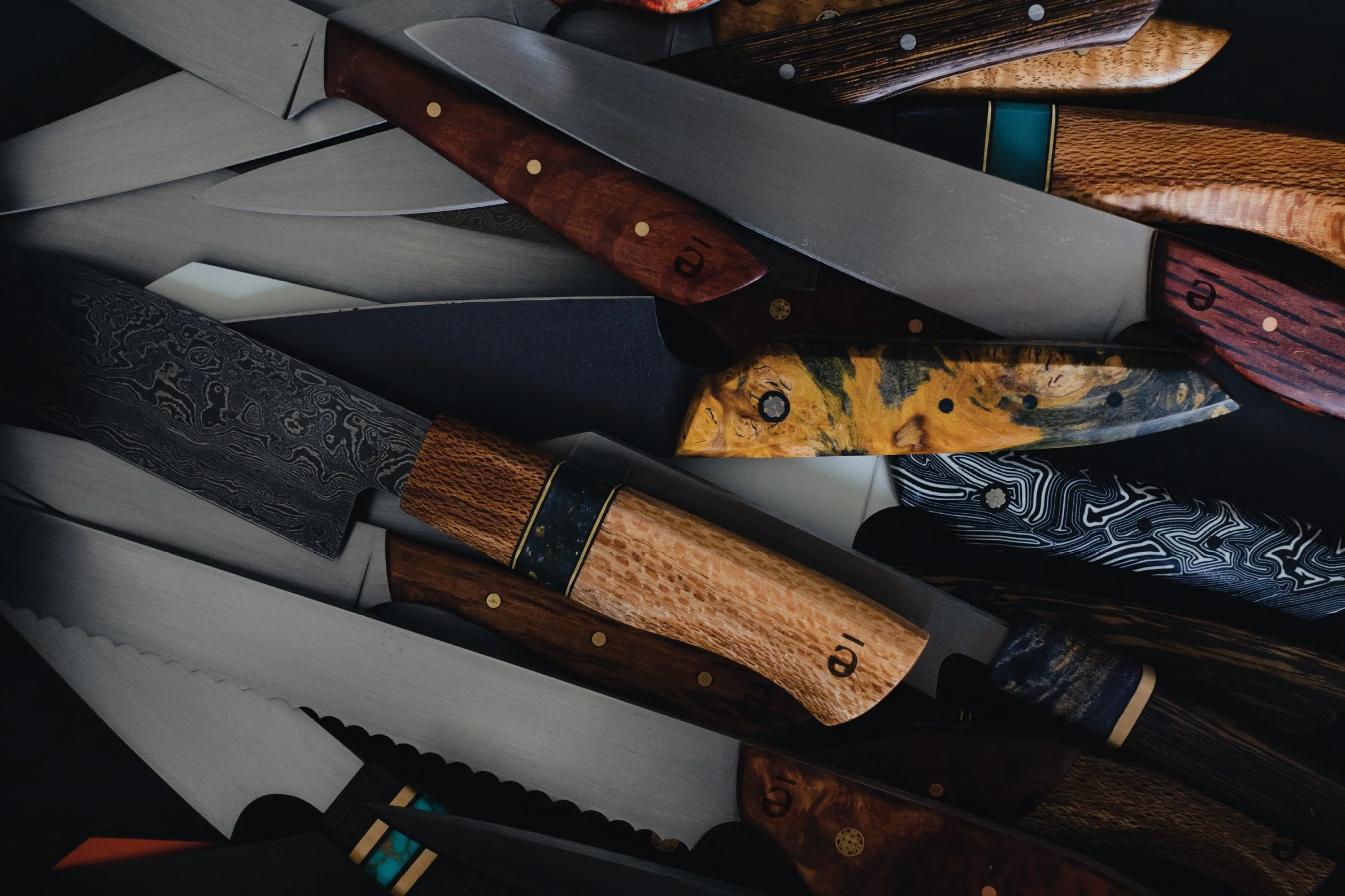 A collection of various knives with distinct wooden and patterned handles, featuring different blade styles and finishes, overlapping each other.