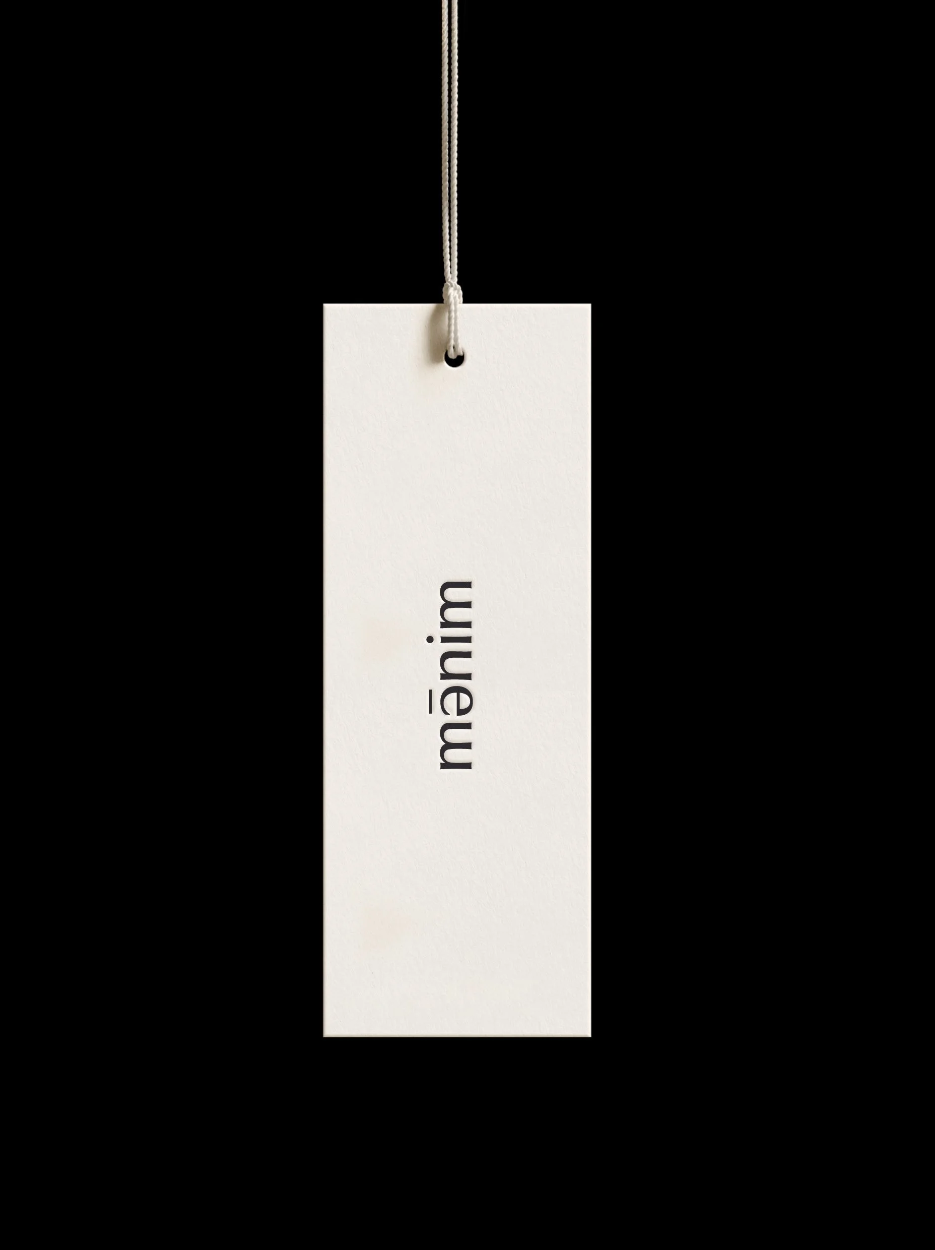 Beige clothing tag with the word 'maim' printed vertically, attached to a string on a black background.