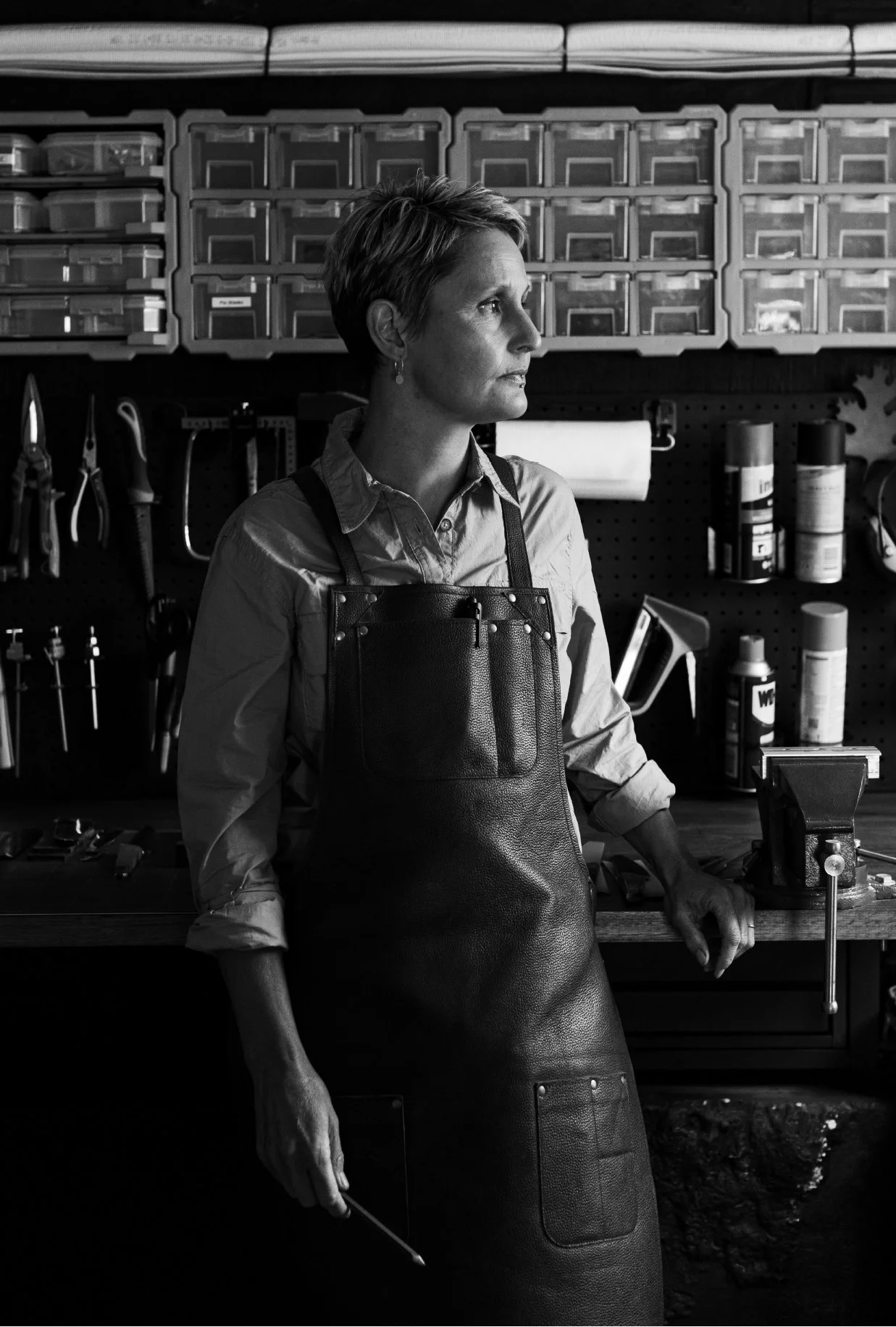 Black and white image of a person in a workshop wearing an apron, standing by a workbench with tools and storage boxes on the wall.