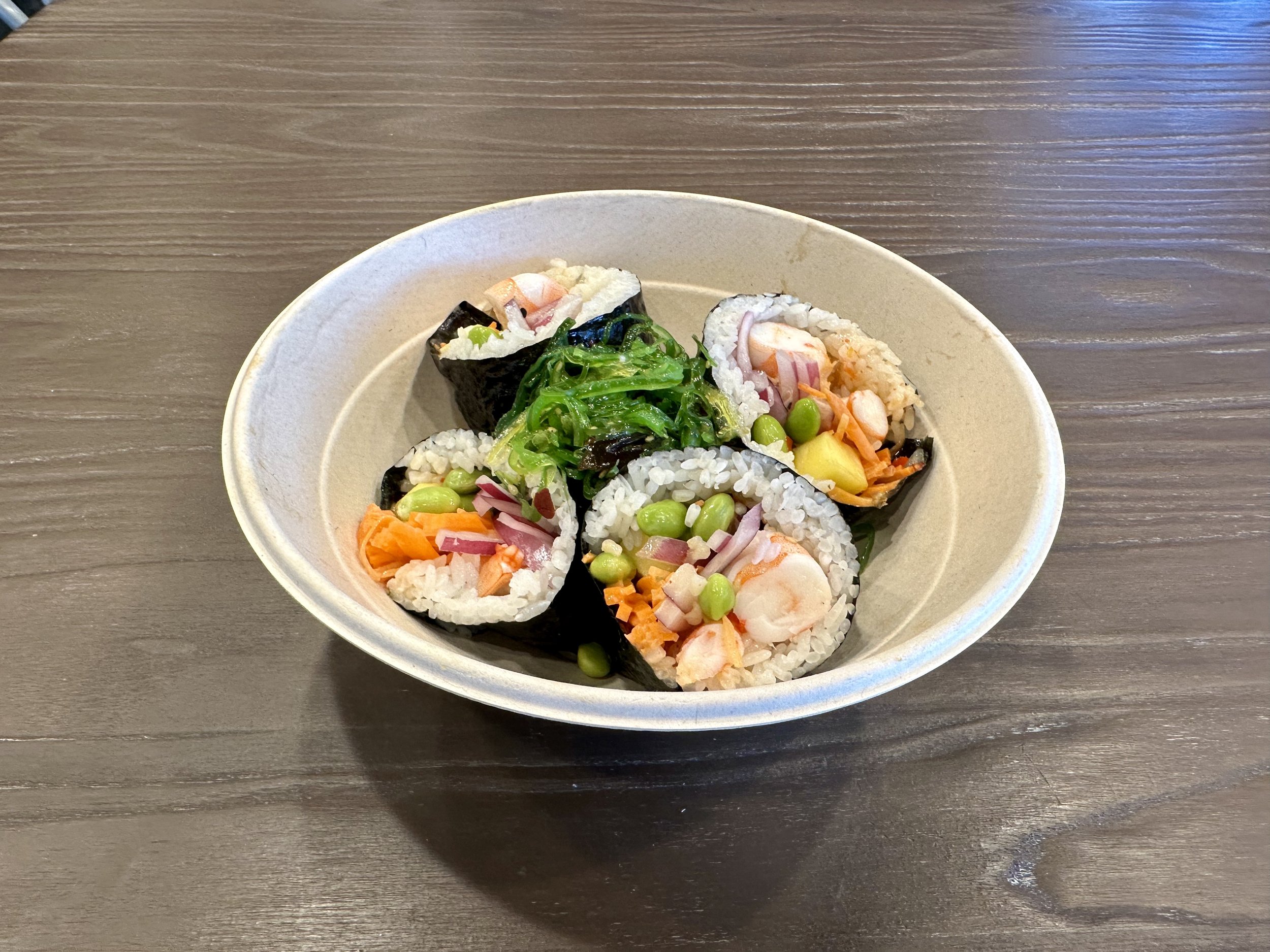 A bowl of assorted sushi rolls with rice, seaweed, shrimp, vegetables, and seaweed salad on a wooden table.