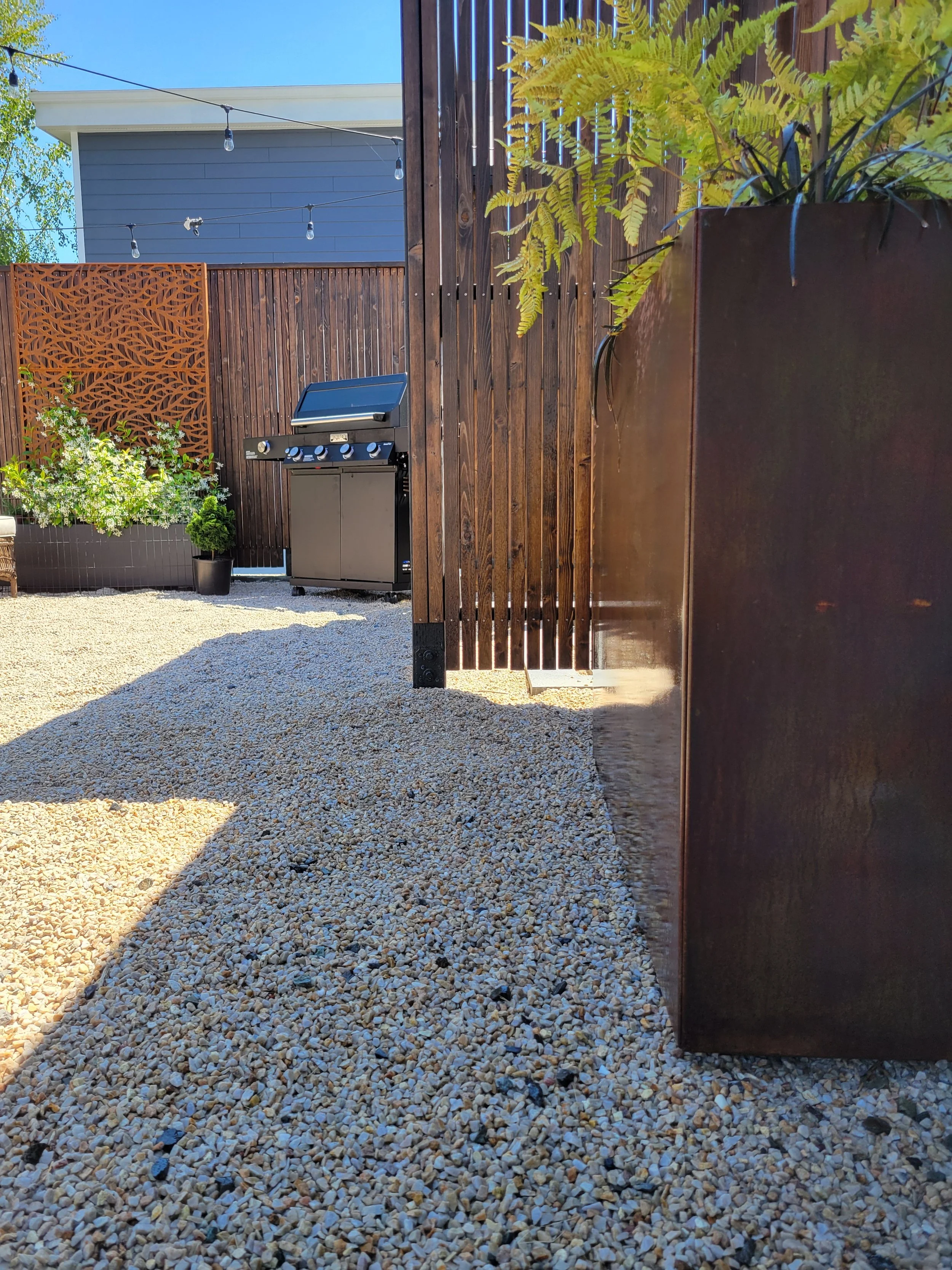 A backyard patio with gravel ground, a wooden privacy fence, and a blue house in the background. There is a barbecue grill, some potted plants, and a corten steel planter box with low-maintenance plantings. String lights hang overhead.