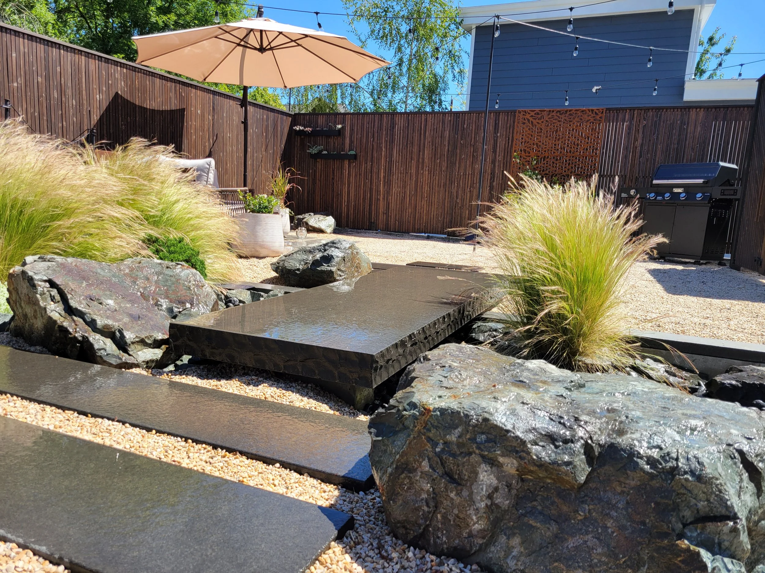 Backyard patio with a dry creek water feature, tall grasses, a stone bridge, rocks, a grill, an umbrella, string lights, and a custom wooden fence.