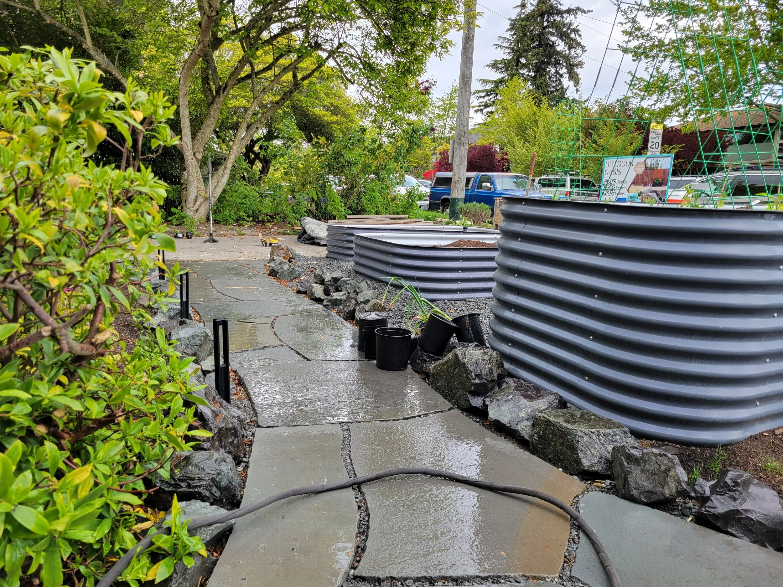 Wet stone pathway in a garden with green shrubs on the left and planters with rocks and soil on the right. In the background, there are trees, a few cars, and outdoor garden supplies.