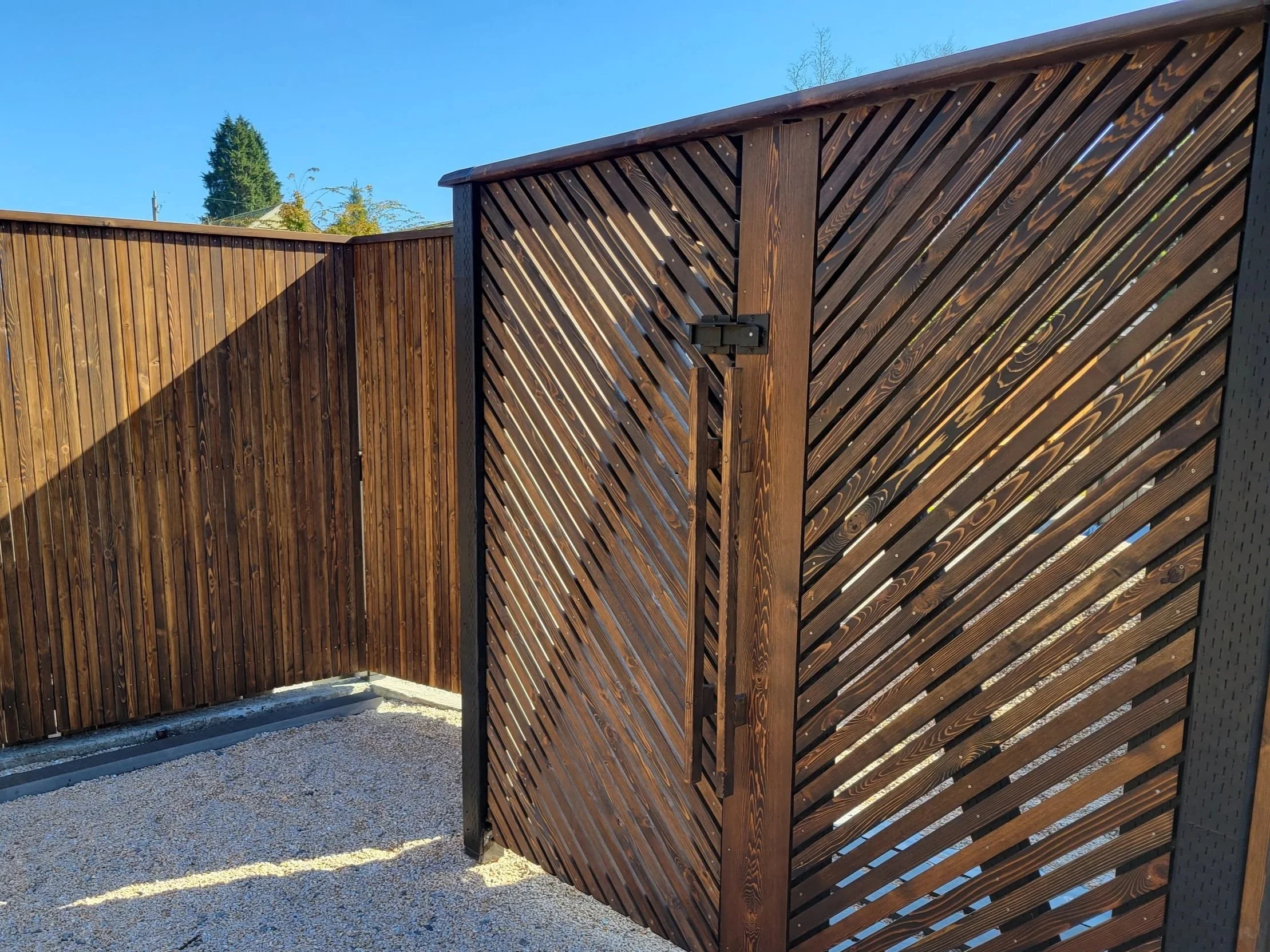 A wooden privacy screen with diagonal slats, secured with a latch, stands outdoors against a backdrop of a clear blue sky, trees, and a fence.