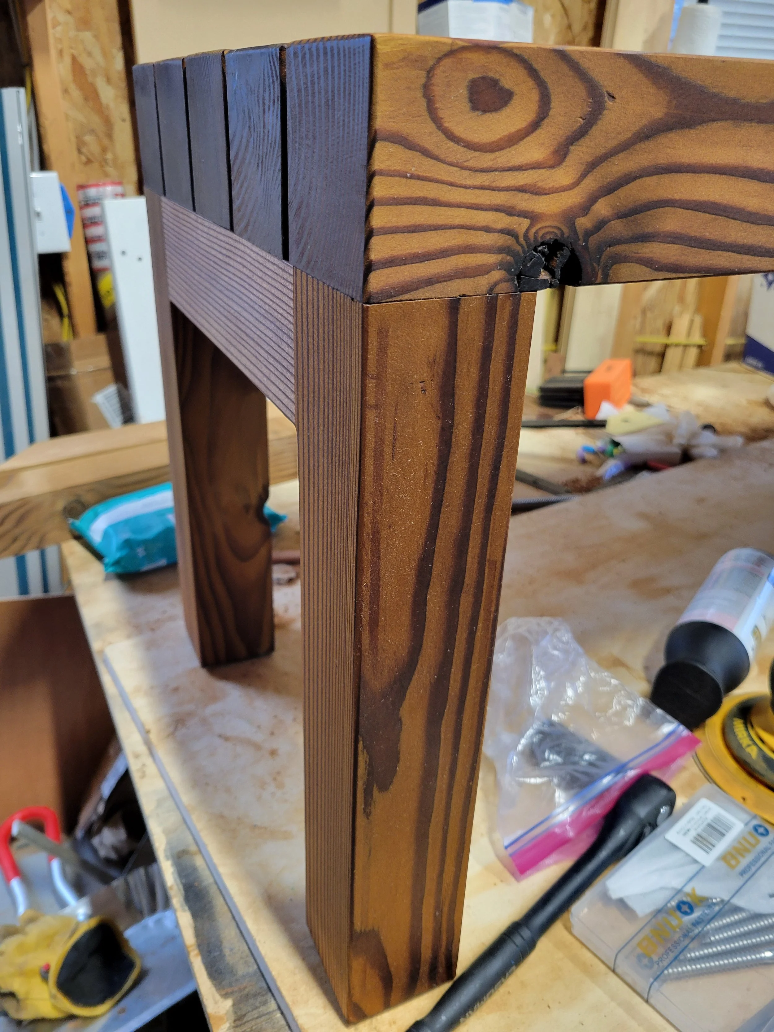 Close-up of a corner of a wooden table or shelf with dark and light stained wooden planks, on a cluttered workbench with tools and supplies in the background.