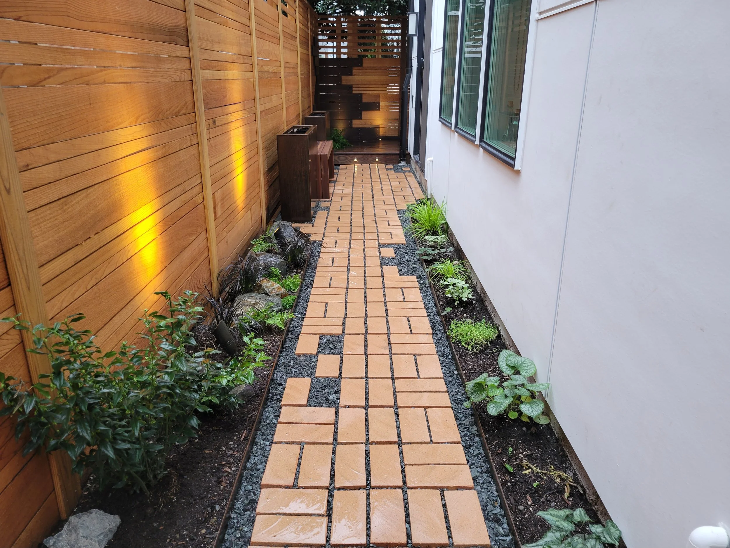 A narrow outdoor walkway with terra cotta bricks, flanked by green, white-flowering plants on the left and a white wall with windows on the right, leading to an artistic woodworking facade and wooden fence, and deck at the end with soft lighting.