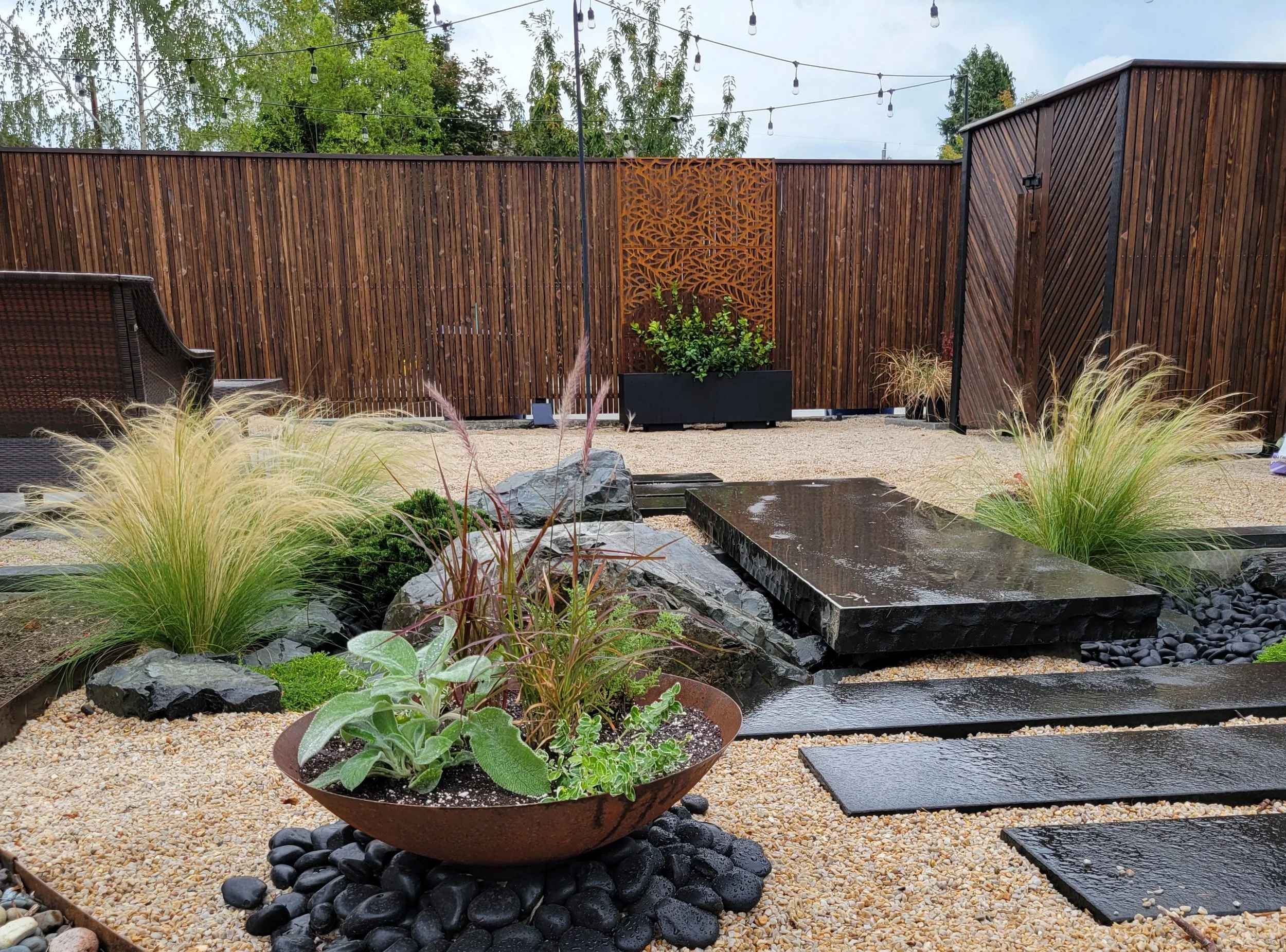 A modern backyard garden with a wooden fence, decorative metal panel, and string lights overhead. Features include plants in a brown dish, black gravel, black stepping stones, and a water feature with rocks.