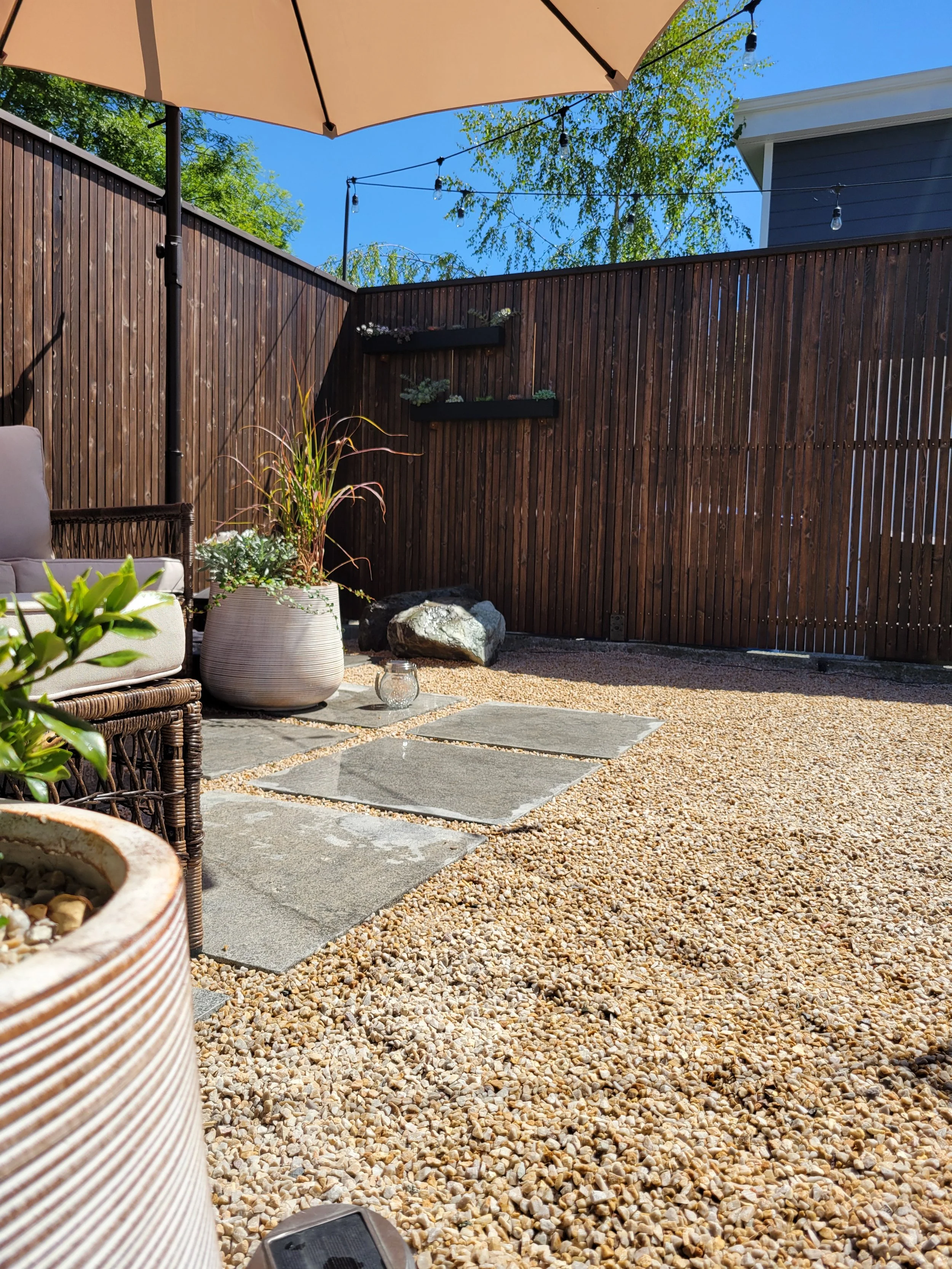 A backyard patio with a wooden fence, potted plants, large rocks, a gravel ground, a stone path, a large umbrella, string lights, and a blue sky with trees in the background.