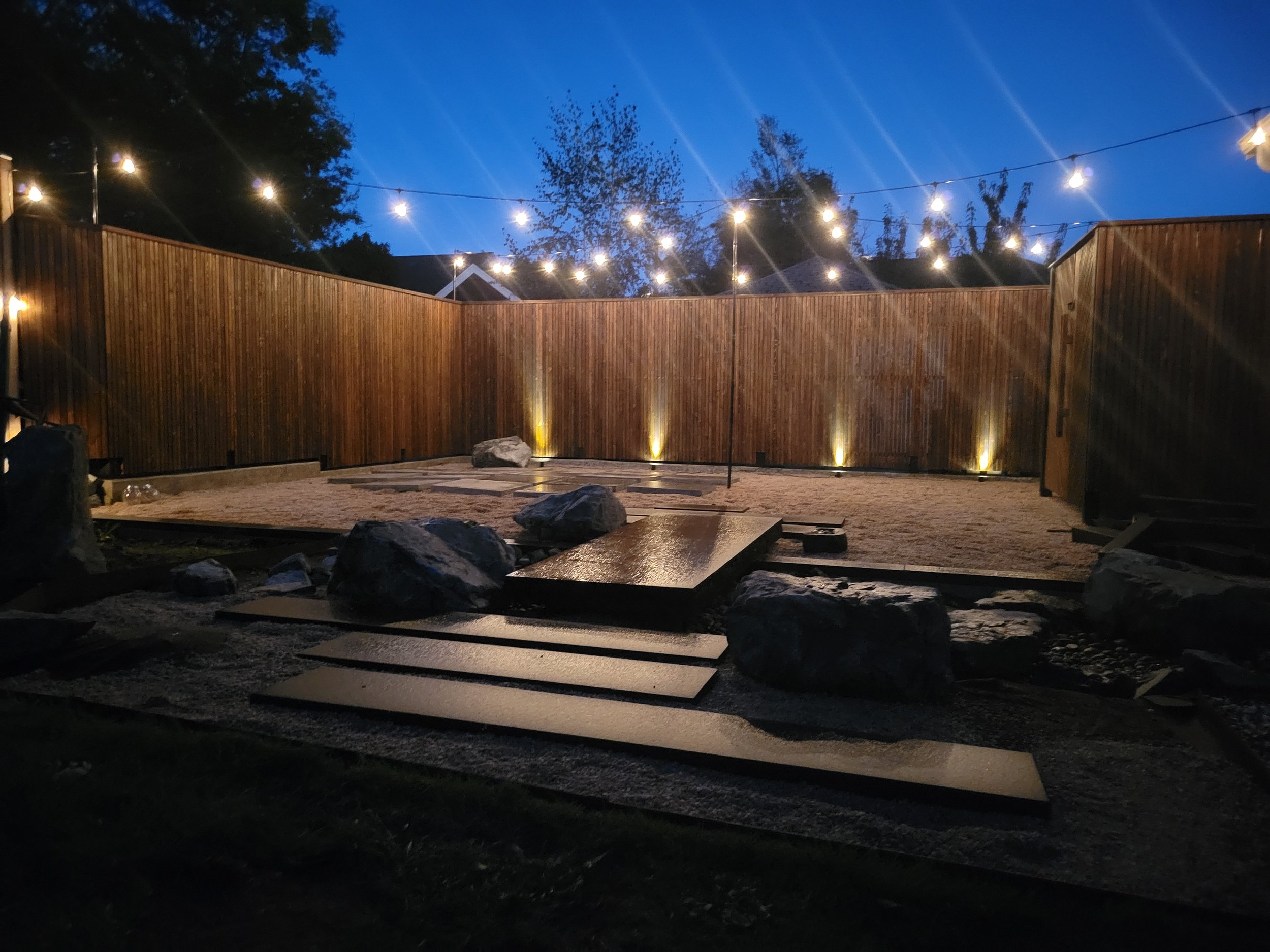 Backyard with wooden privacy fence, string lights overhead, rocks, and a gravel area, possibly a garden or outdoor entertainment space at dusk.