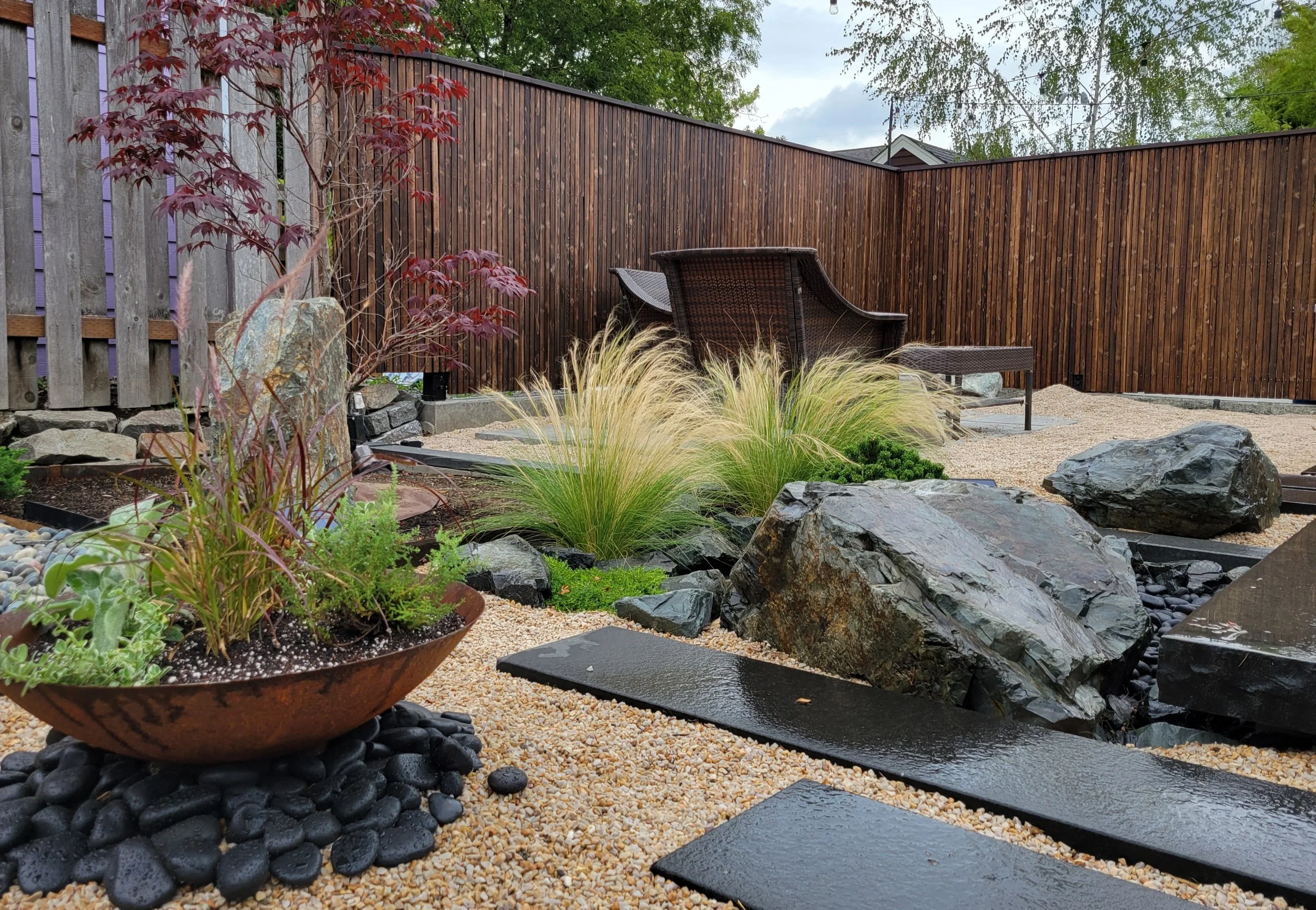 A landscaped backyard with a wooden privacy fence, ornamental plants, rocks, and stepping stones.