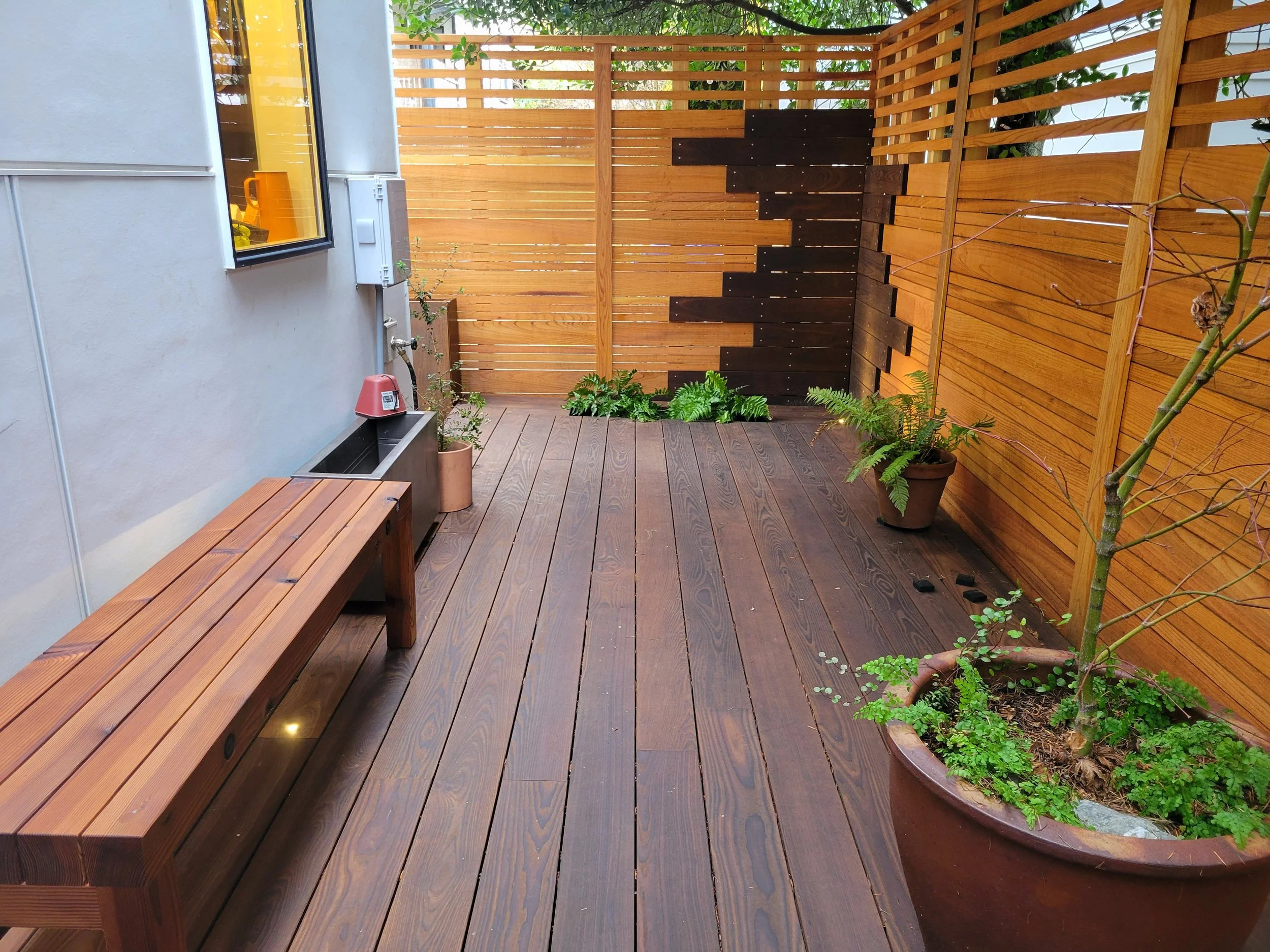 A small patio with wooden flooring, a wooden privacy fence, a potted plant, recessed planter box with ferns, and a bench.  The fence has a custom woodworking facade and uplighting and the deck bench also has uplighting.