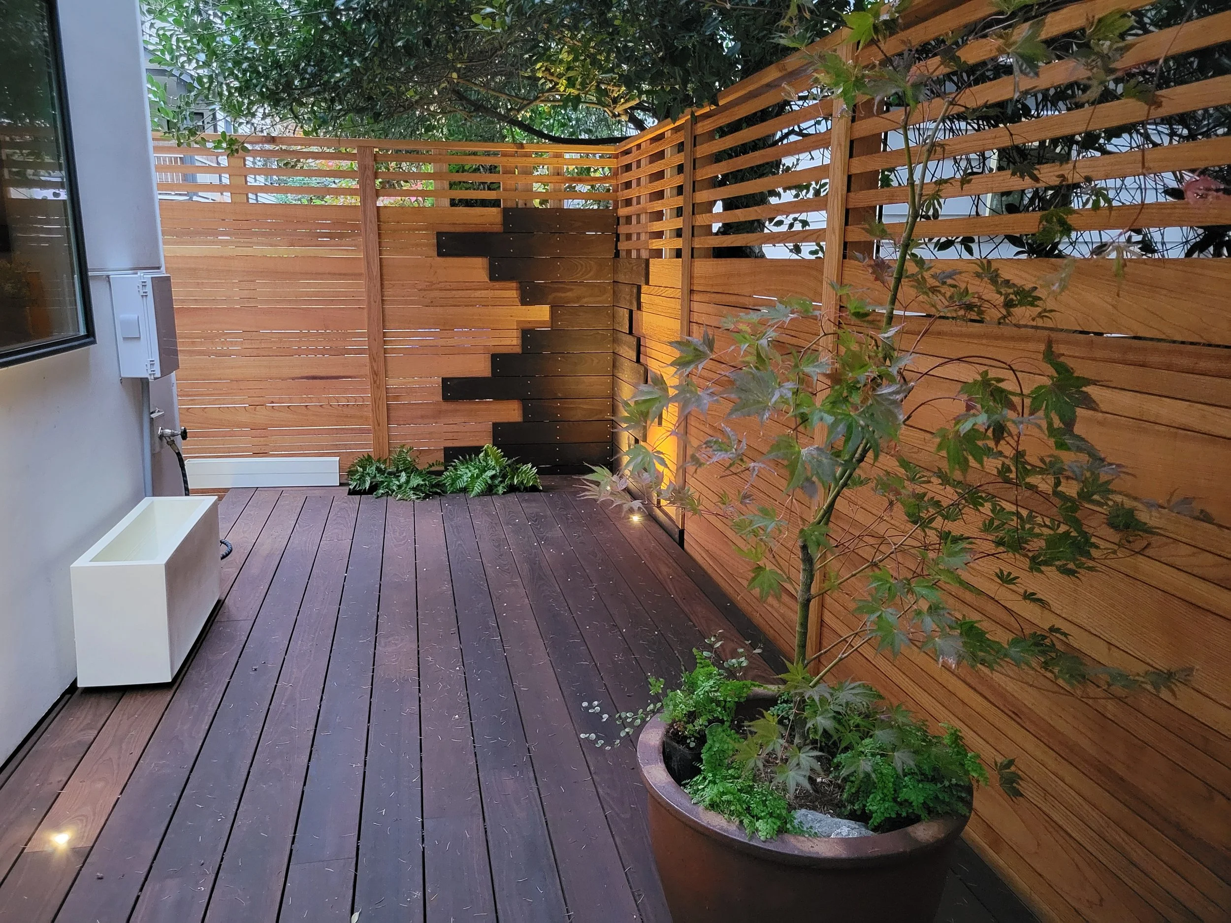 A small outdoor patio with wooden decking, a wooden privacy fence, potted plants including a small tree, and outdoor lighting.