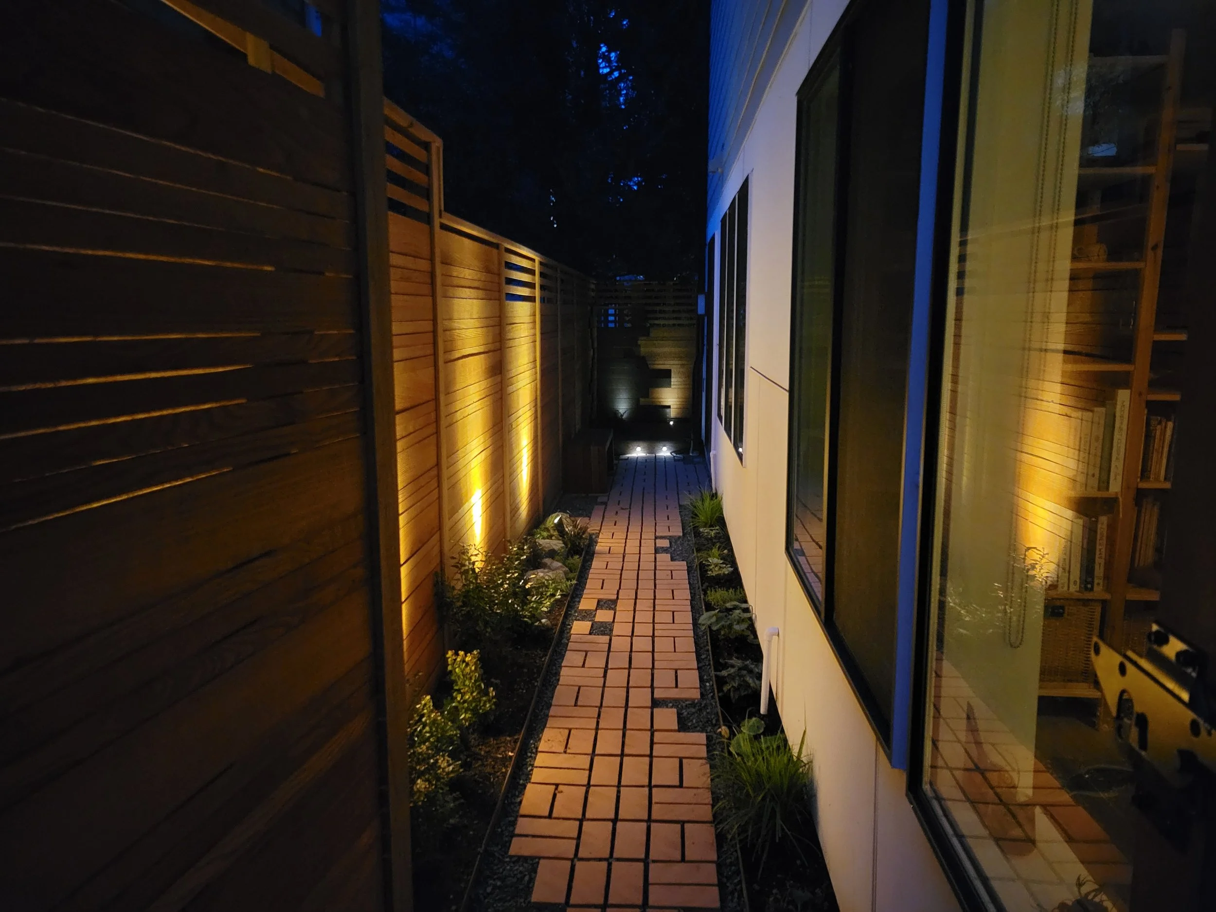 Night view of a narrow brick pathway between a wooden fence and a house with large windows, illuminated by outdoor lighting.