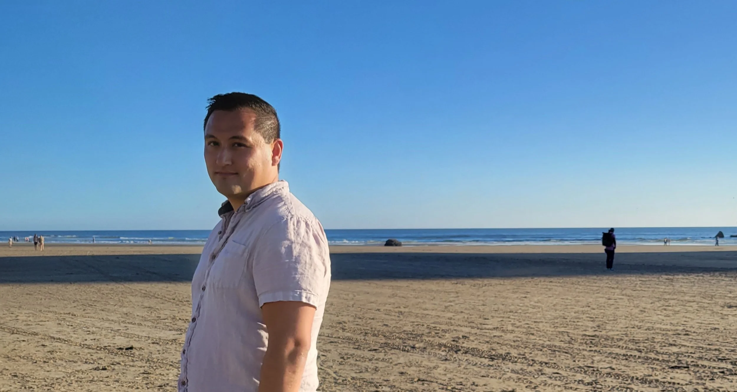 The owner of Outdoor Oasis standing on a sandy beach near the ocean on a sunny day, with a blue sky overhead and people in the distance, some walking and some sitting, enjoying the beach scene.