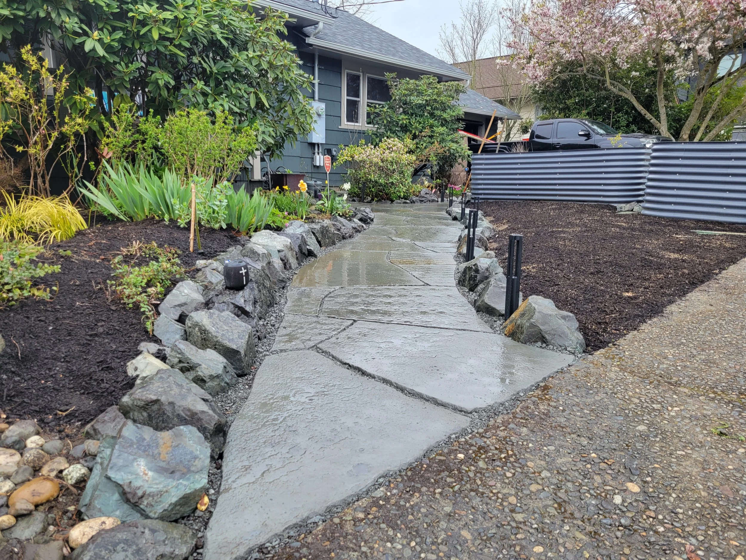 A winding blue-stone garden pathway and natural stone rockery meant to mimic the effect of a river.  Landscape lighting lines either side of the pathway.  Before the planting beds mature.