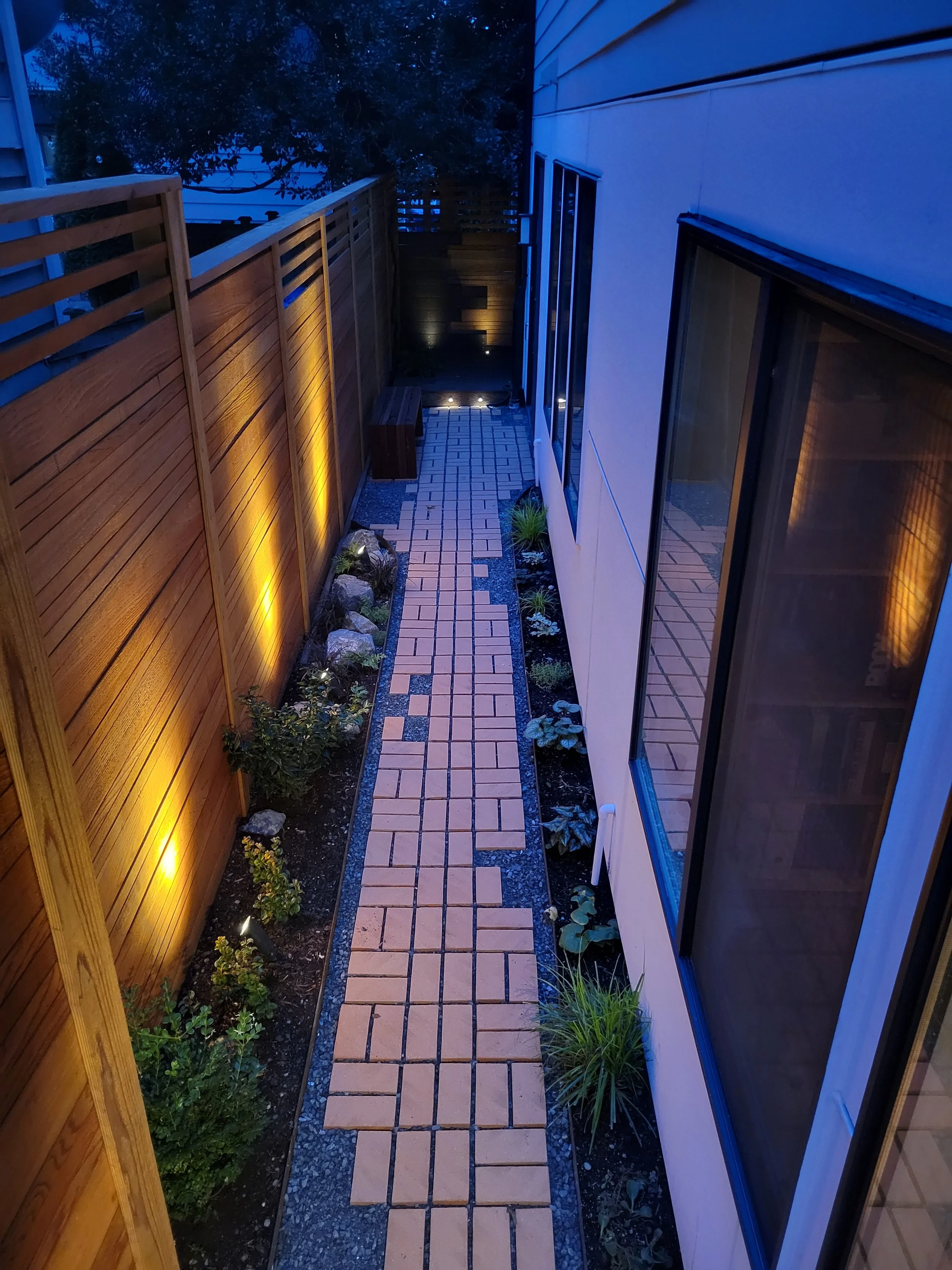 A narrow backyard pathway at dusk with brick paving, lit by warm landscape lighting along a wooden fence on the left and a white house with large windows on the right.