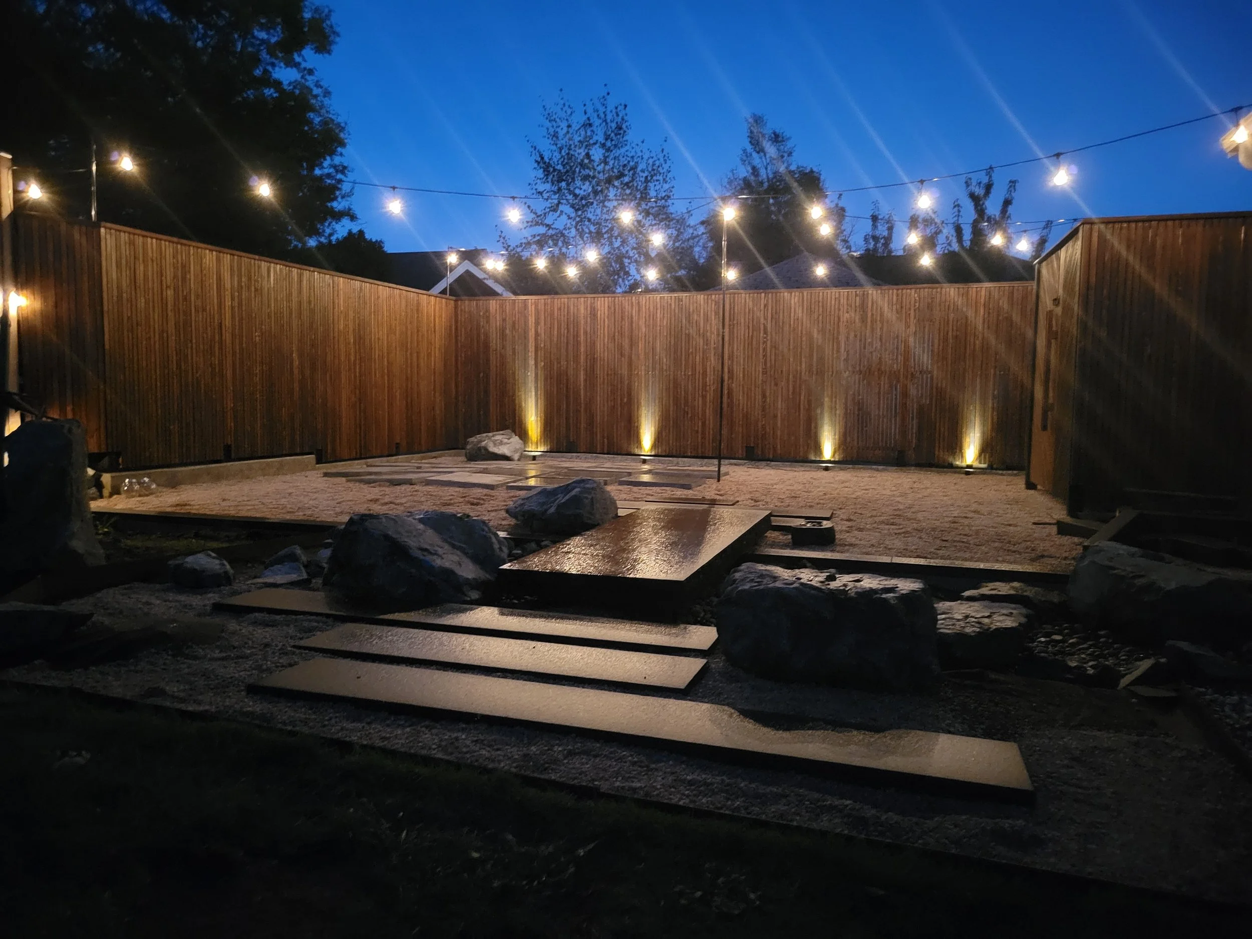 Nighttime backyard scene with string lights hanging above a wooden fence, some rocks and paving stones on the ground, and tall trees silhouetted against the dark blue sky.