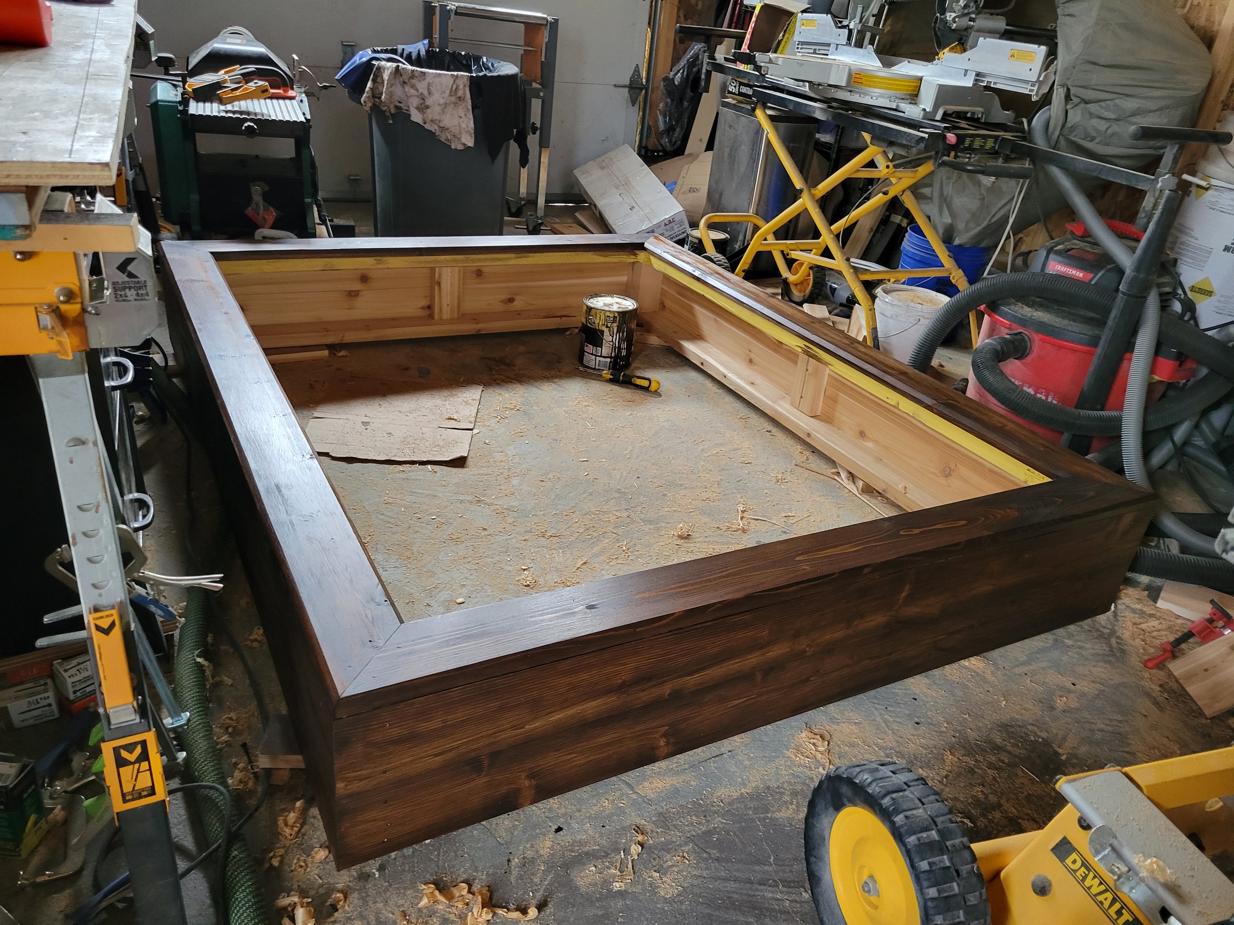 A woodworking workshop with a partially assembled wooden frame, various tools, and equipment like saws, vacuum, and hand tools scattered around.