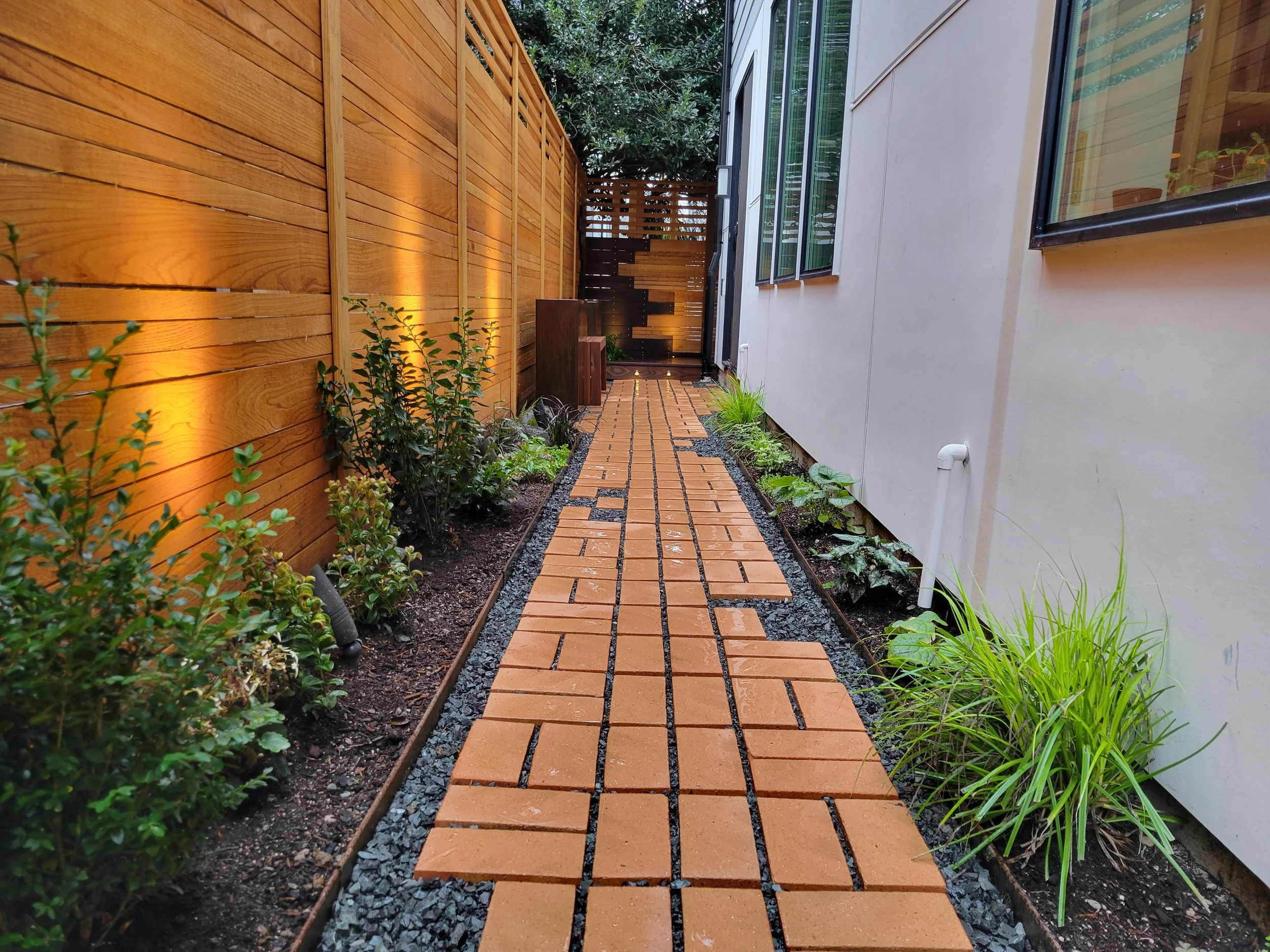 A narrow outdoor walkway with terra cotta bricks, flanked by green, white-flowering plants on the left and a white wall with windows on the right, leading to an artistic woodworking facade and wooden fence, and deck at the end with soft lighting.
