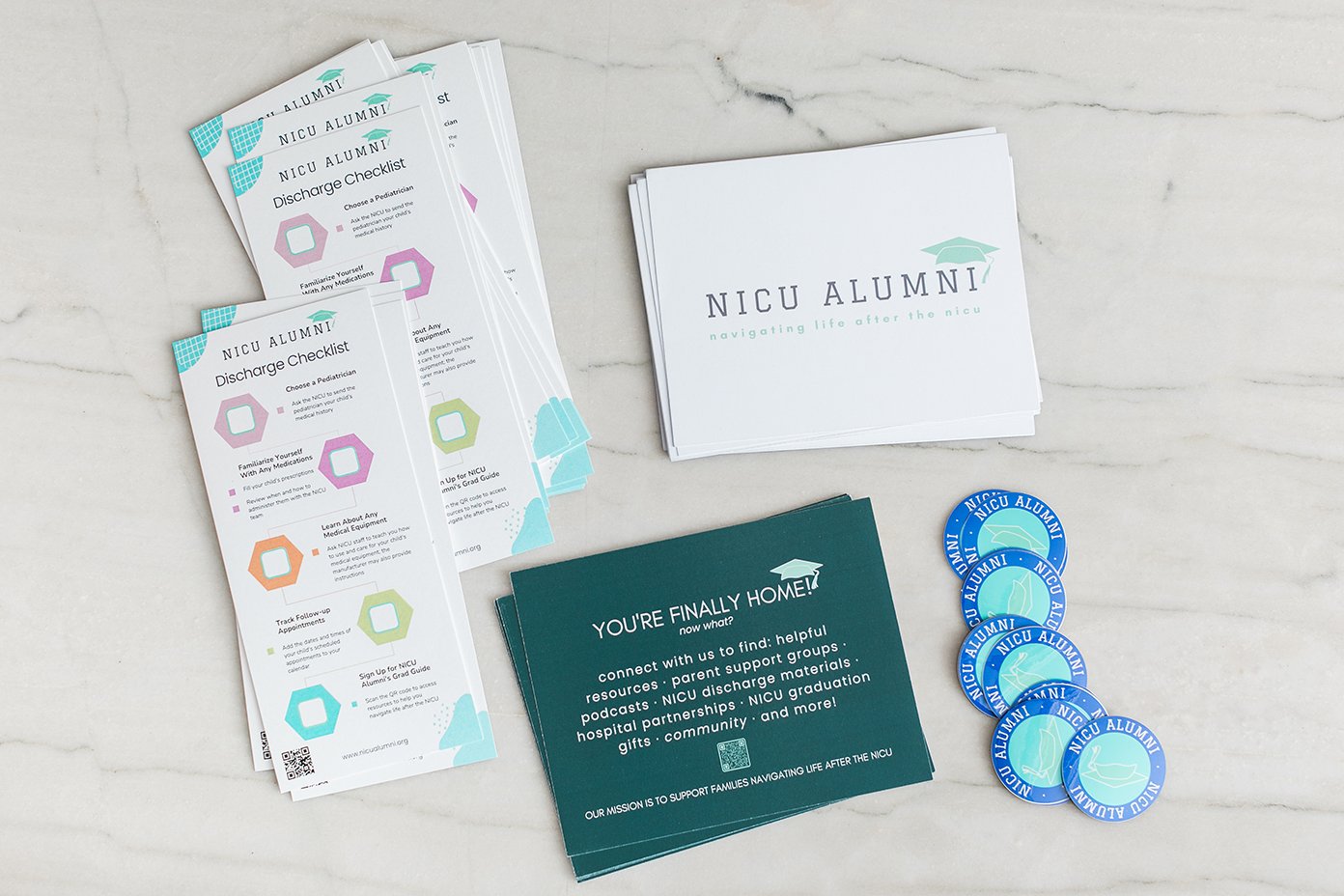 NICU Alumni informational materials including discharge checklist pamphlets, a stack of cards with NICU Alumni logo, and four NICU Alumni wristbands on a white marble surface.