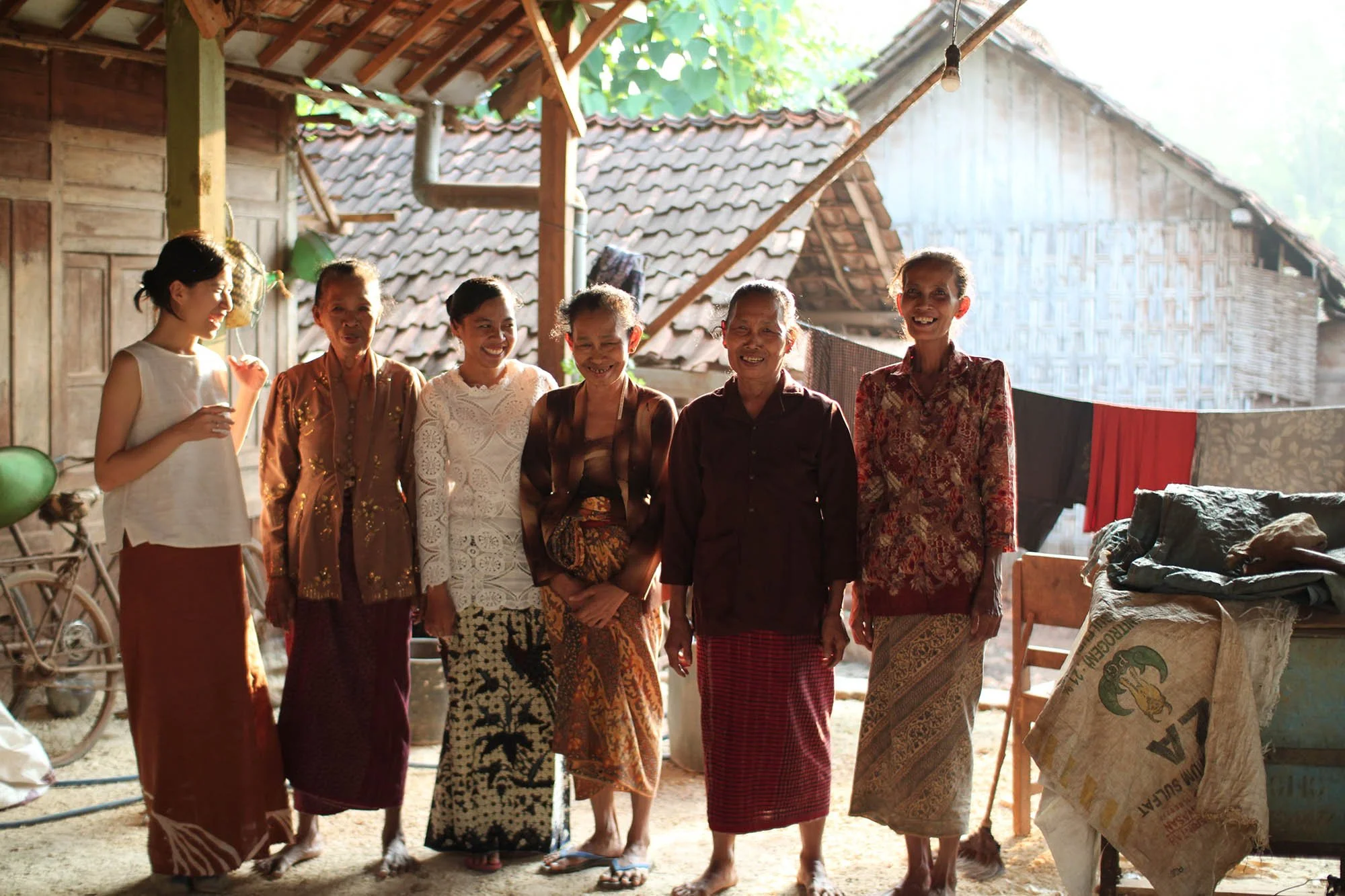 A group of six women standing together, smiling, in front of rustic buildings with tile roofs. They are wearing traditional clothing and appear to be in an outdoor setting with some laundry hanging and a bicycle visible in the background.