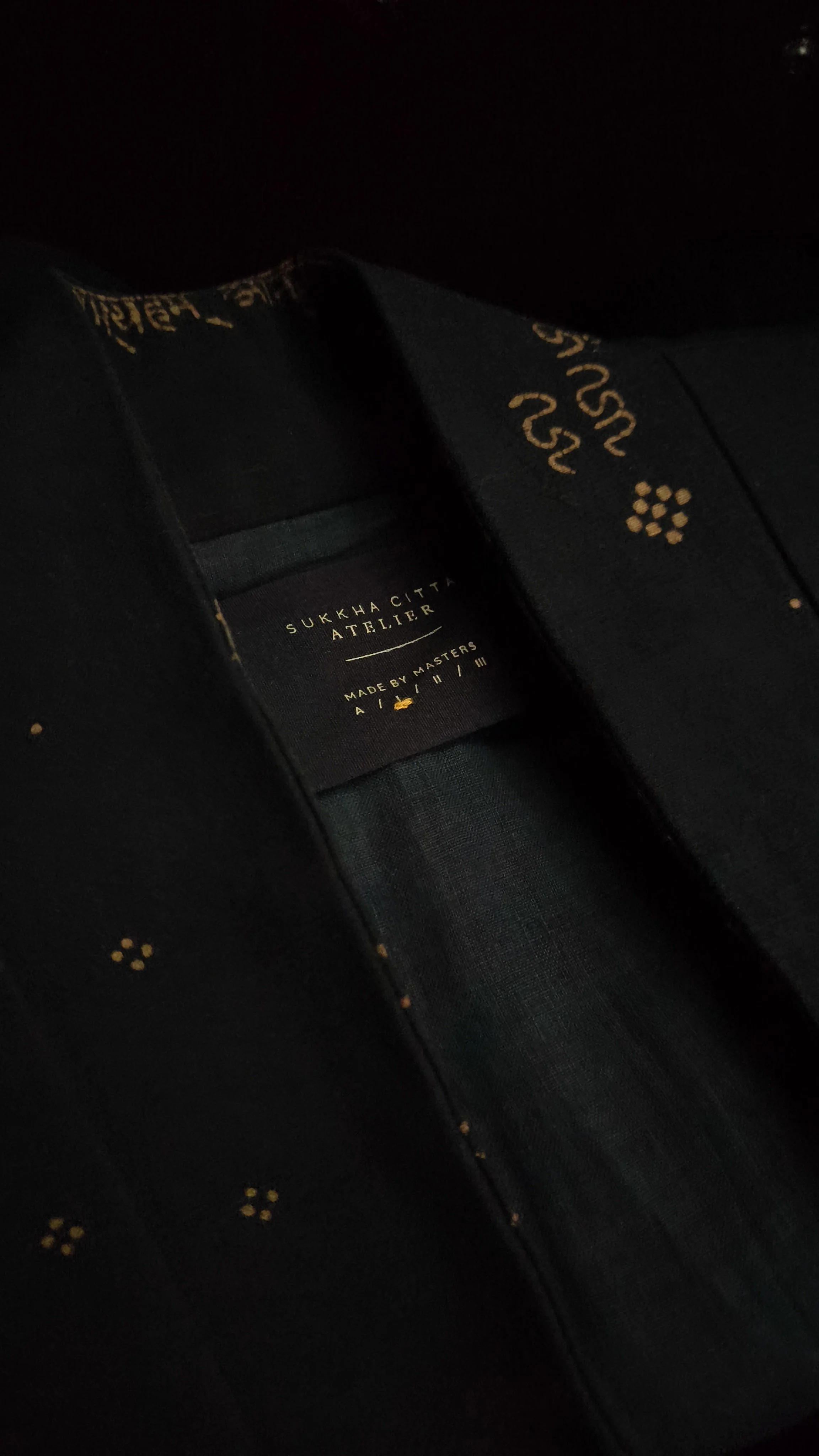 Close-up of a garment label inside a dark fabric with gold patterns, showing text "Sukkhacitta Atelier" and "Made by Masters."
