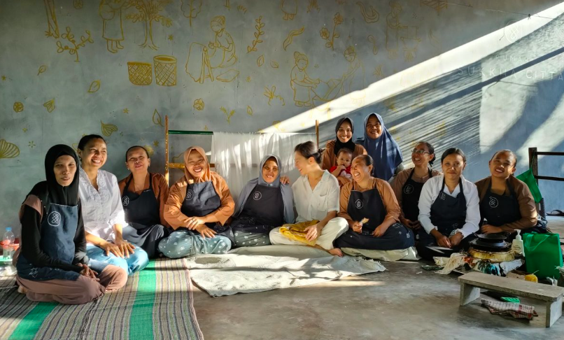 Group of women sitting on mats, smiling at the camera, wearing aprons, in a room with wall art.