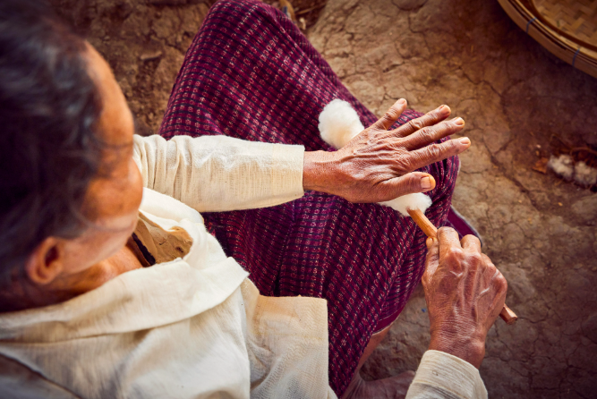 Person spinning cotton thread by hand, wearing a textured maroon skirt and white shirt, seated on brown ground.