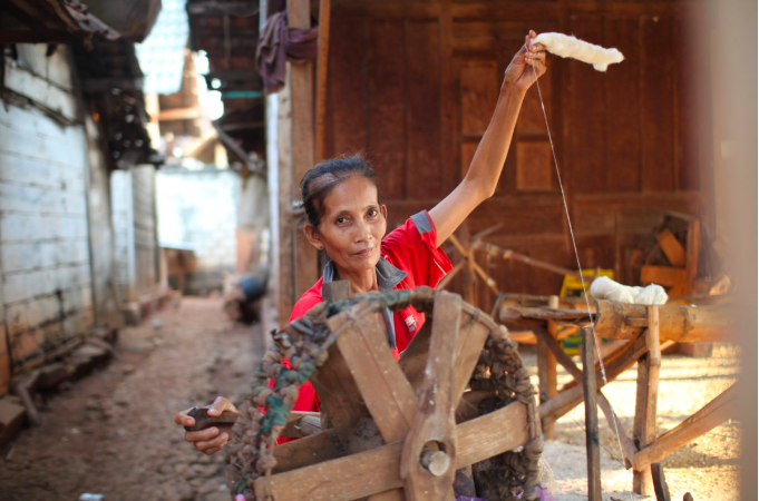 A woman operating a traditional spinning wheel to spin thread from fiber, in an outdoor setting with rustic surroundings.