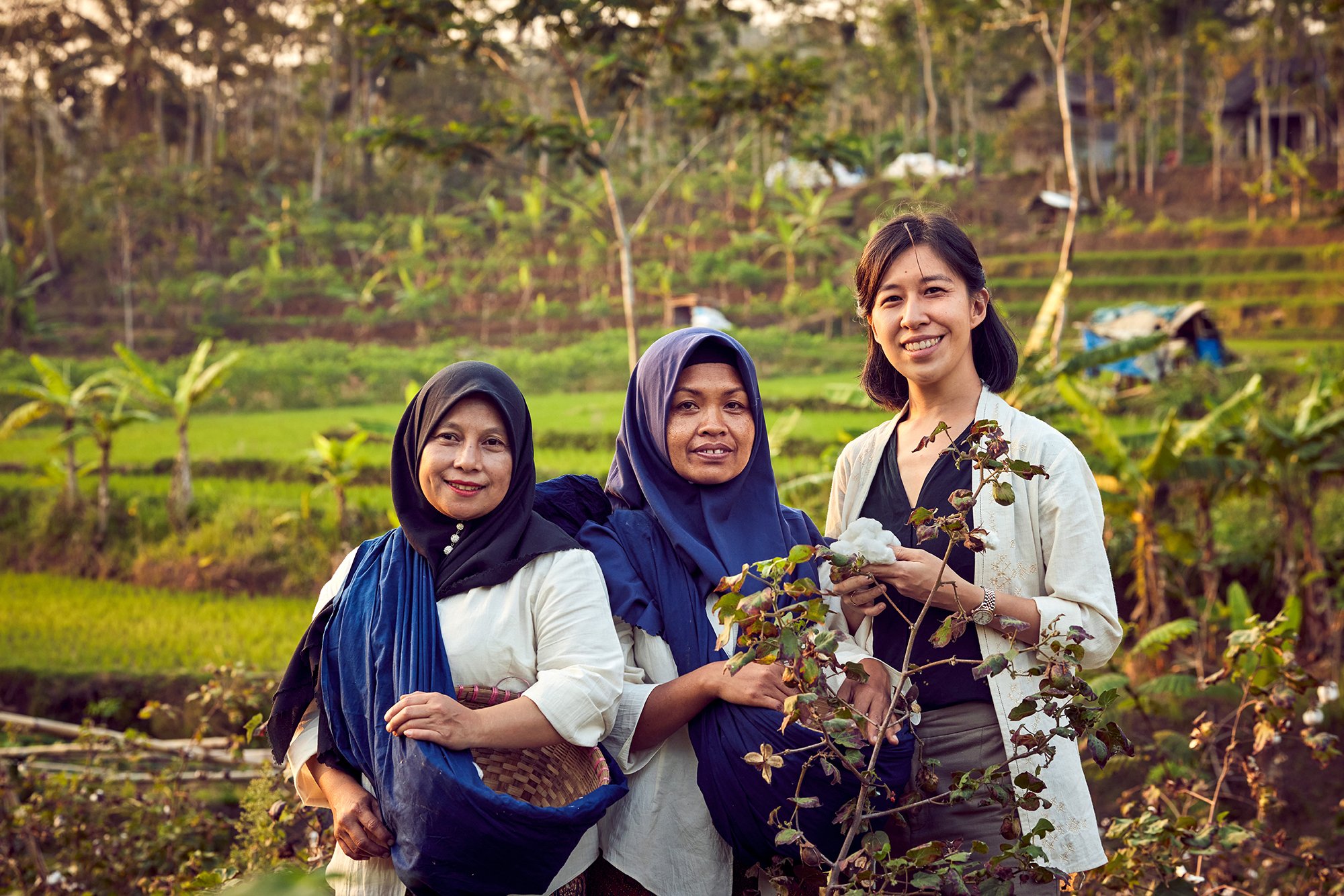 Three women standing together in a lush green agricultural field, smiling at the camera. Two are wearing headscarves, and one is holding a plant.