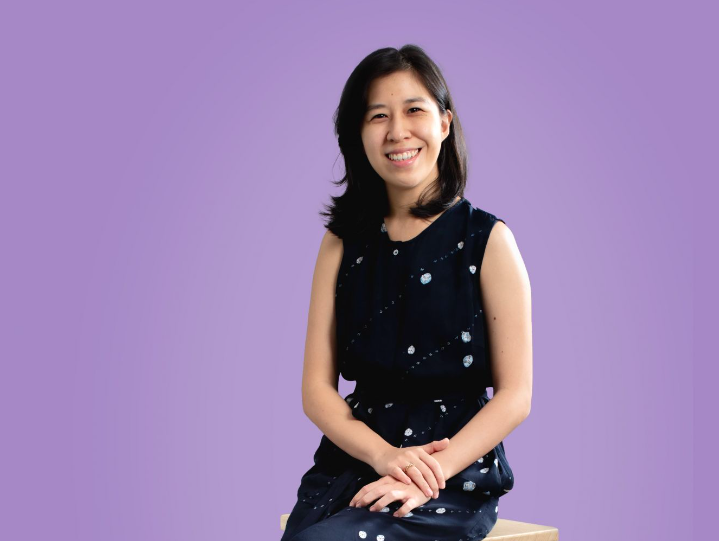 Person in a sleeveless navy dress with patterned dots sitting against a purple background, smiling.